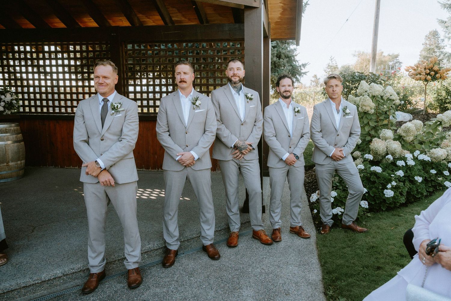 Wedding party members in light gray suits and brown shoes stand casually in a garden setting.