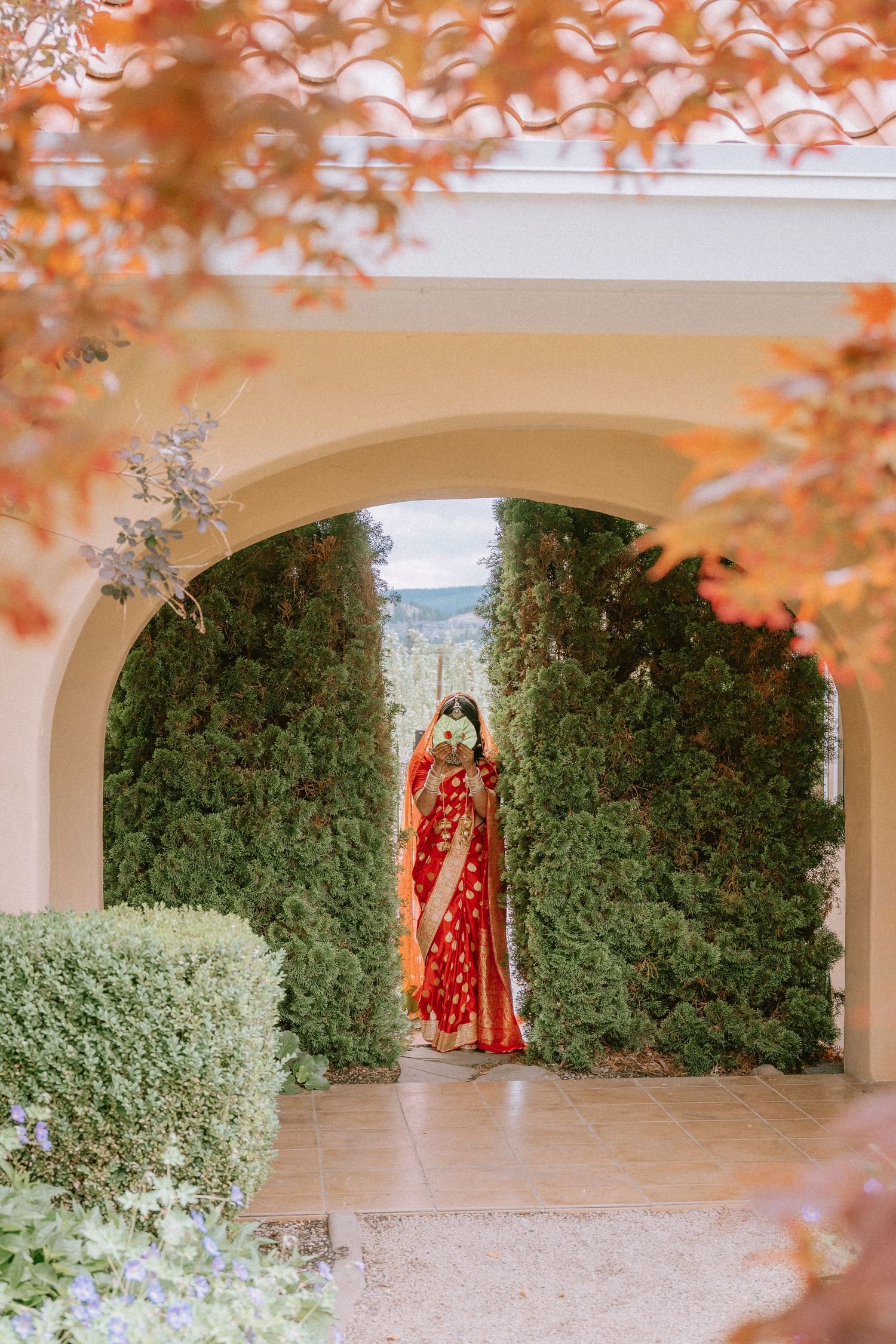 A walkway decorated with autumn foliage and greenery leads to figures in traditional Indian wedding attire.