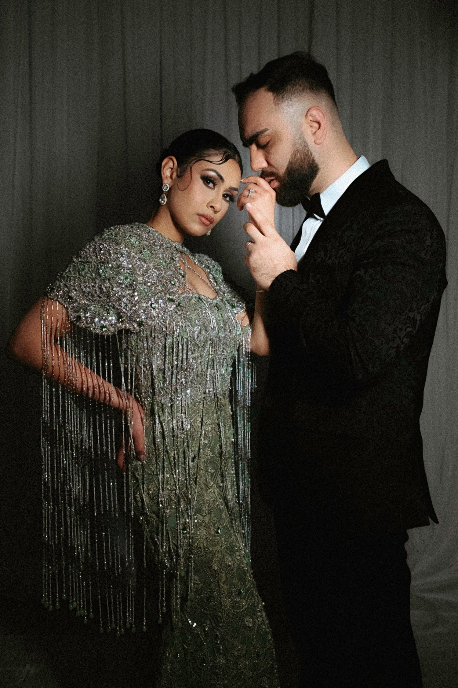 A couple in formal attire poses intimately against dark backdrop, with woman wearing shimmering fringed silver dress.