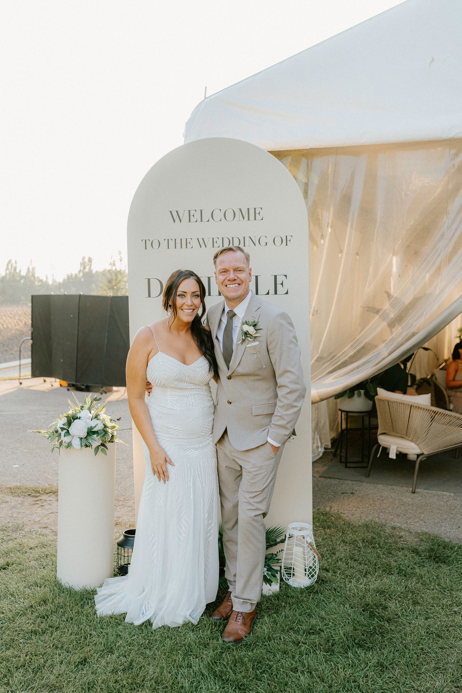 Wedding couple poses by welcome sign outside white tent venue during sunset.