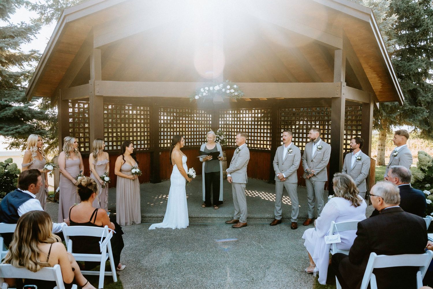 Panoramic view of an outdoor wedding ceremony under a wooden pavilion with guests seated on white chairs.