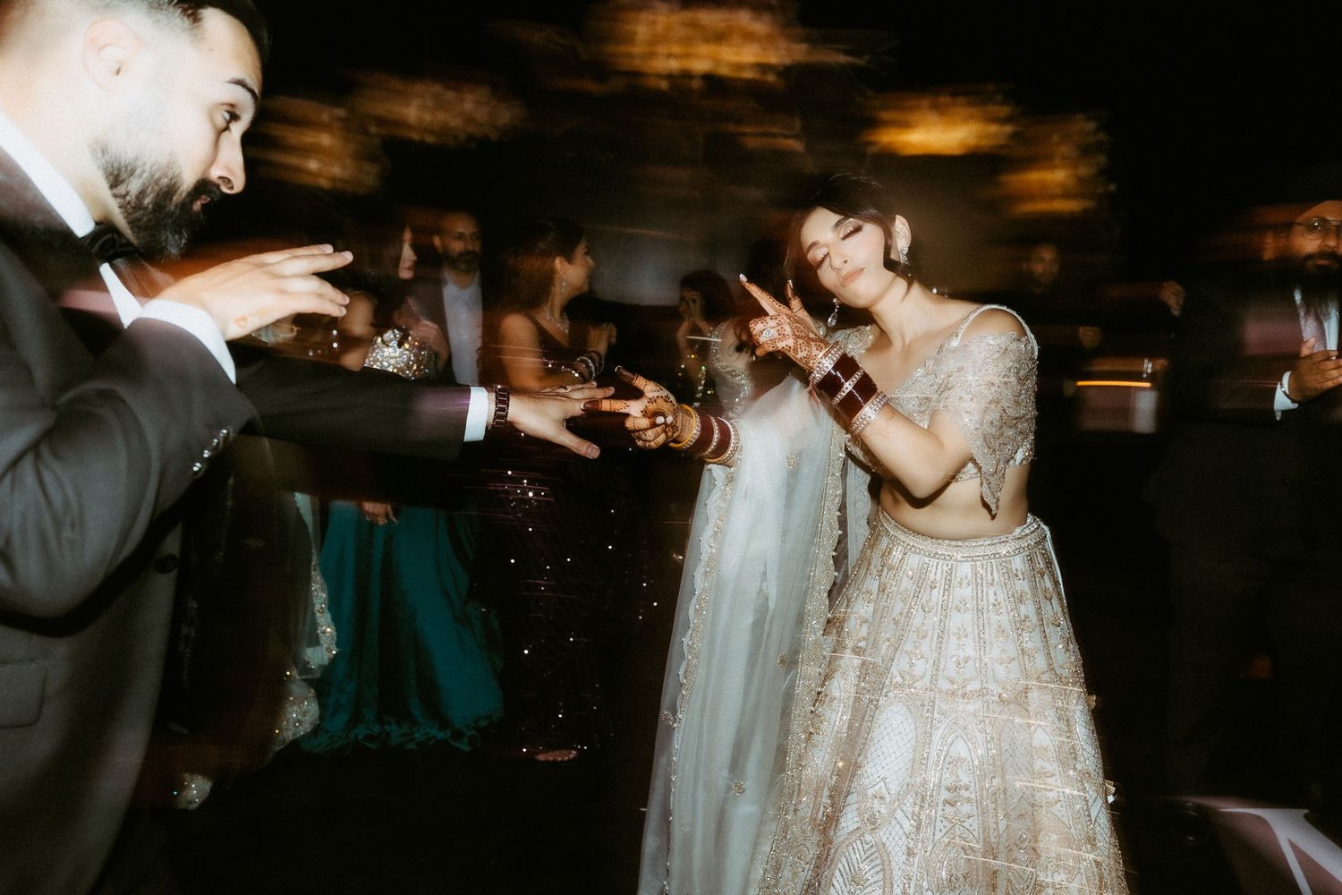 A couple dances joyfully at their wedding reception, with the bride wearing a sparkly white gown in a dimly lit venue.