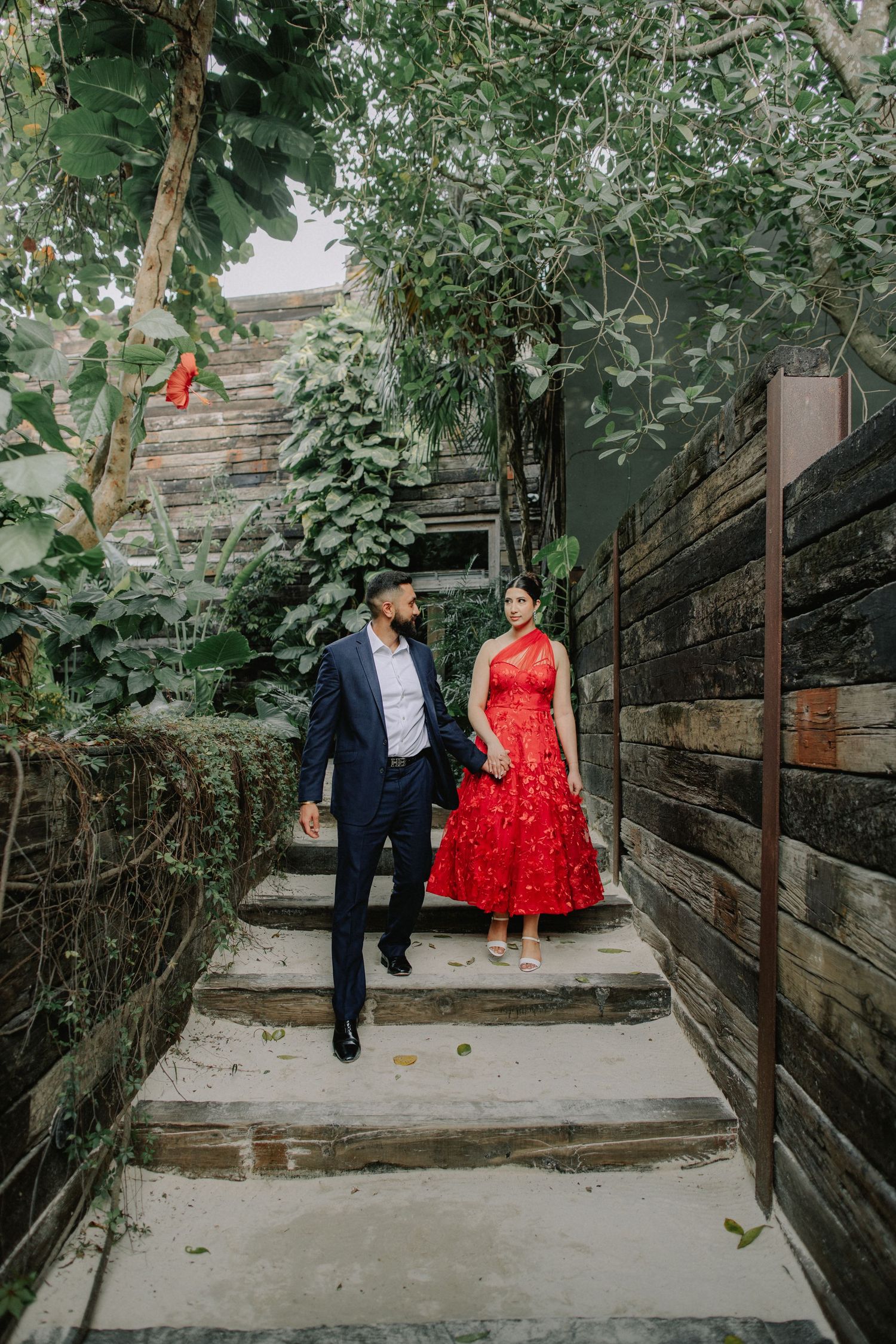 A couple stands on stone steps surrounded by lush greenery, with the person in red wearing a vibrant dress.