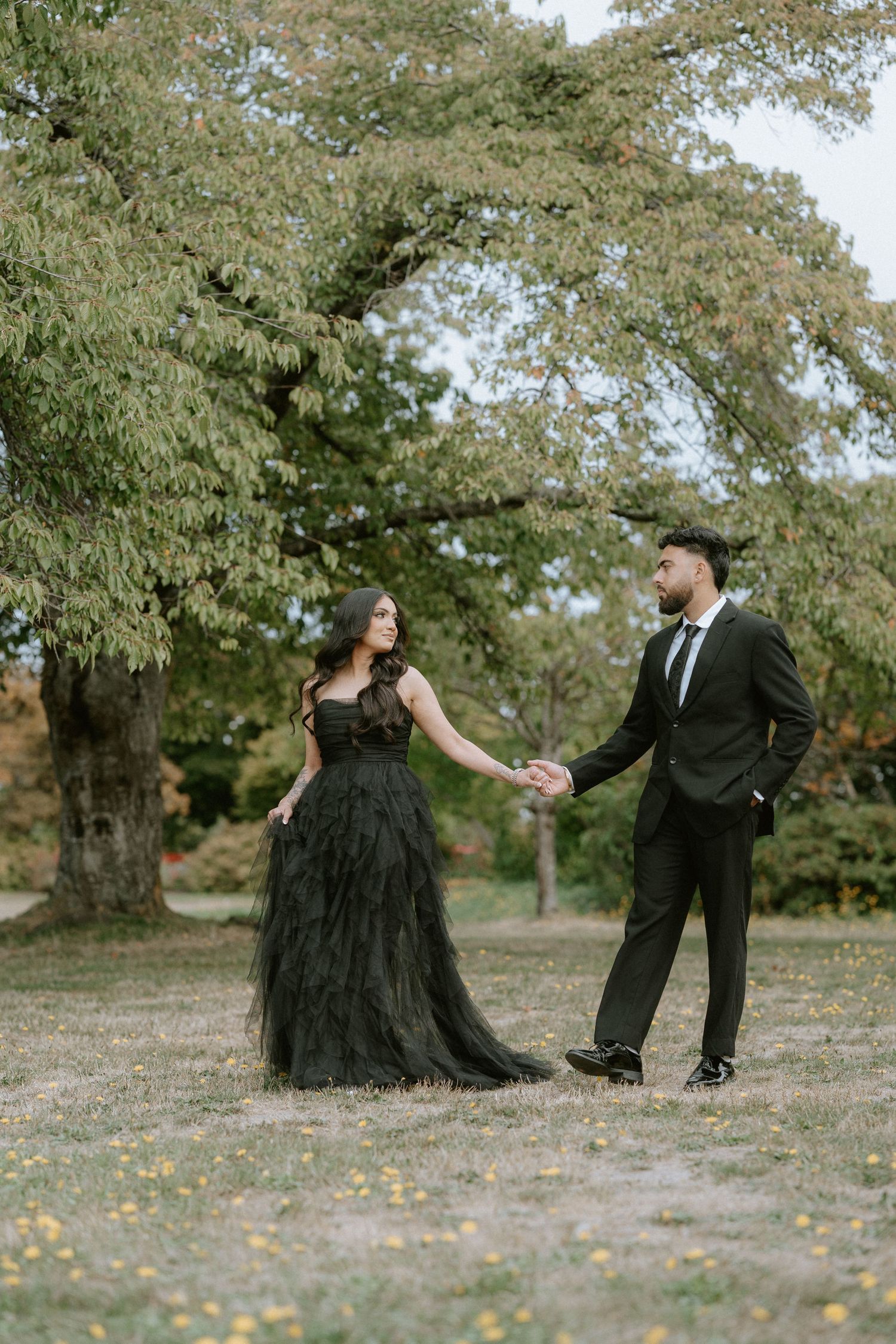 A couple in formal attire dances playfully in a park setting with autumn trees in the background.