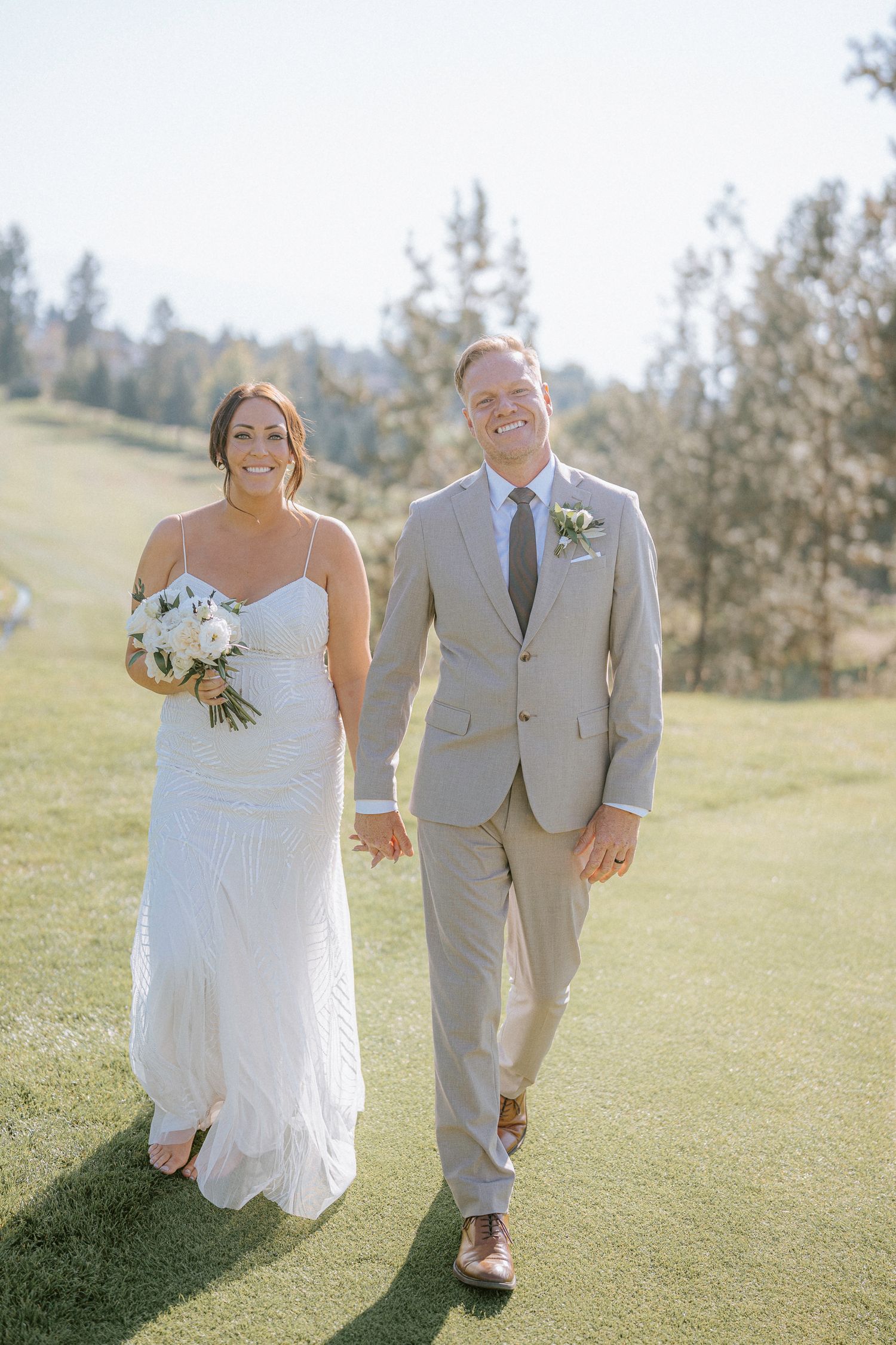 A happy couple walks hand in hand across a sunny hilltop meadow during their wedding celebration.