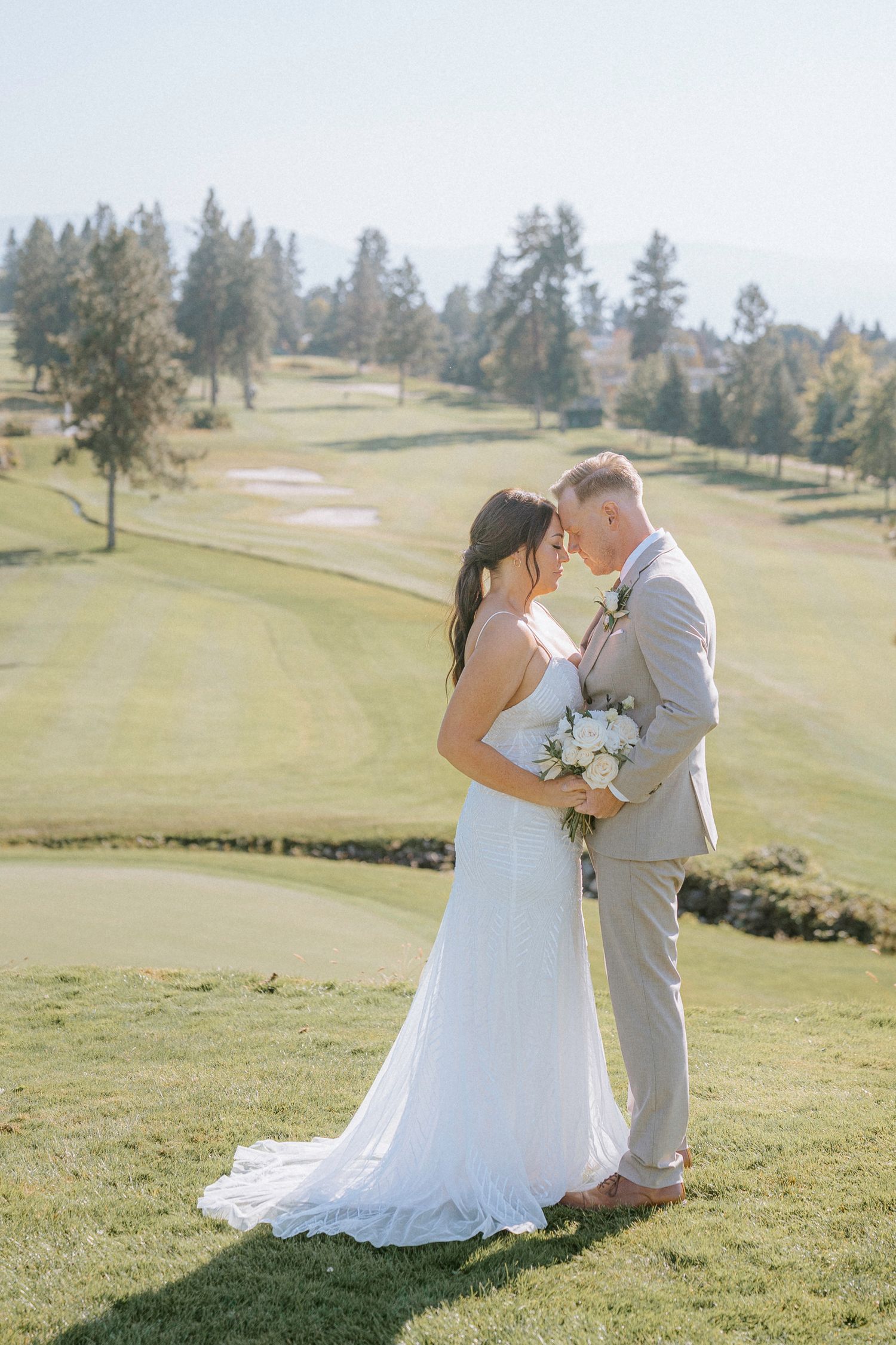 Newlyweds embrace on a scenic golf course with rolling hills and evergreen trees in the background.