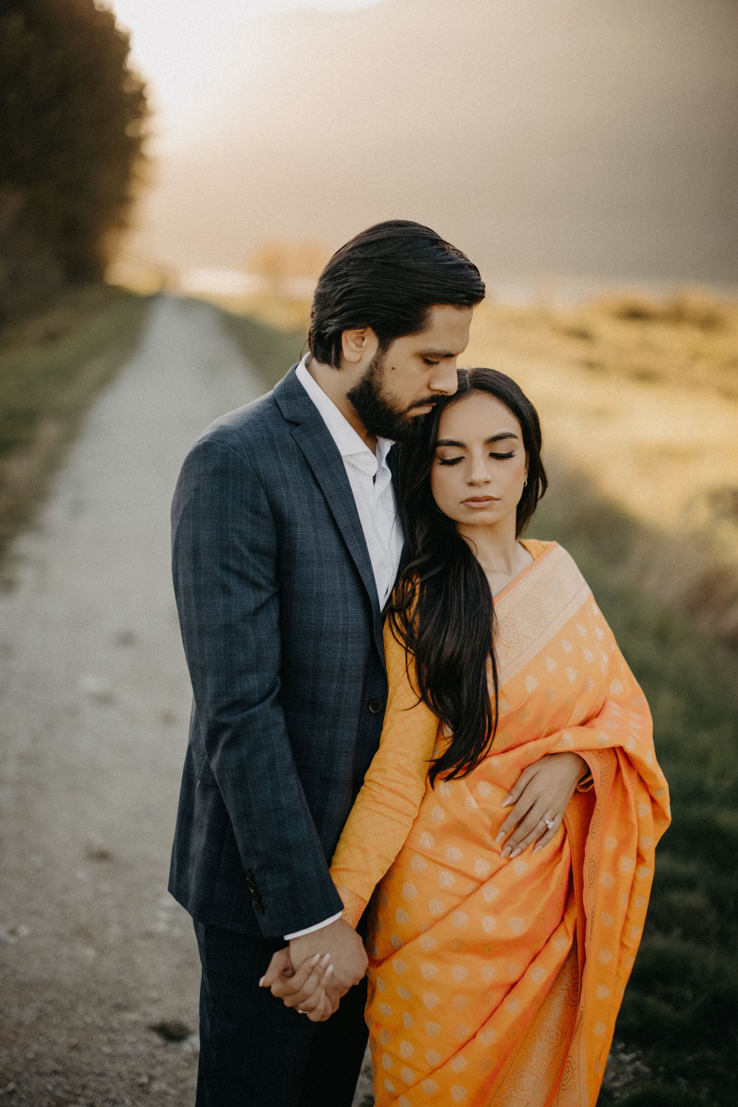 A romantic couple in traditional Indian attire poses together on a dirt path during golden hour.