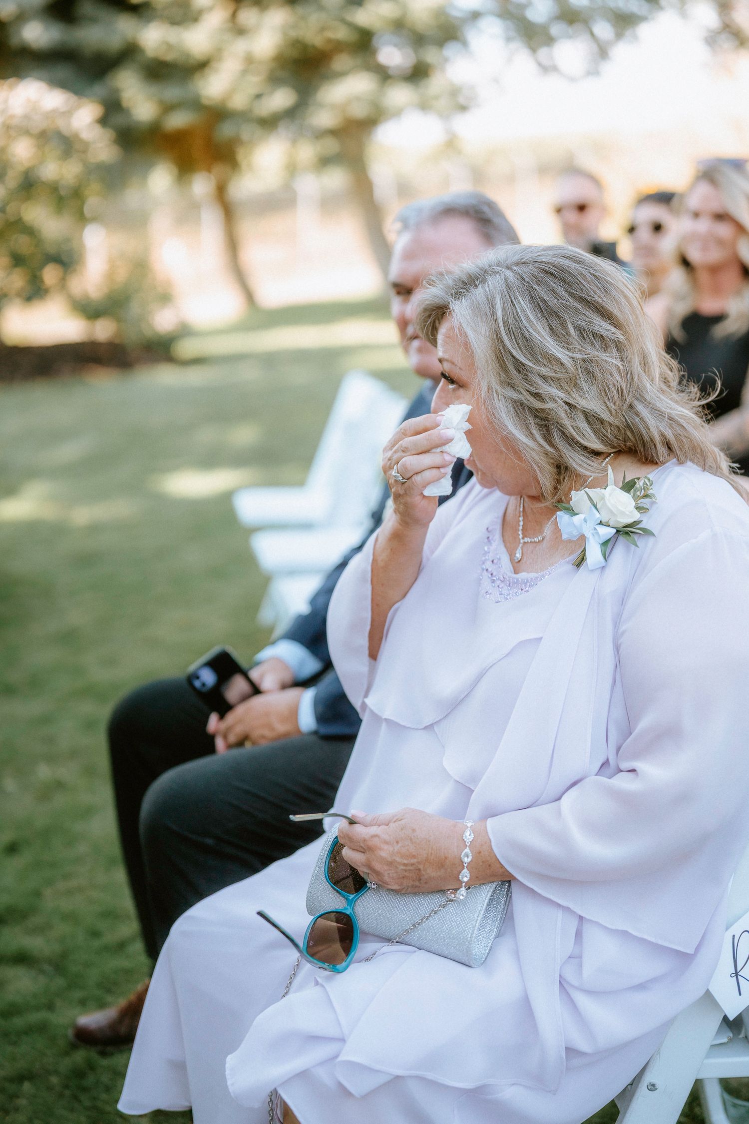 Wedding guests in white attire seated outdoors during an emotional ceremony moment.