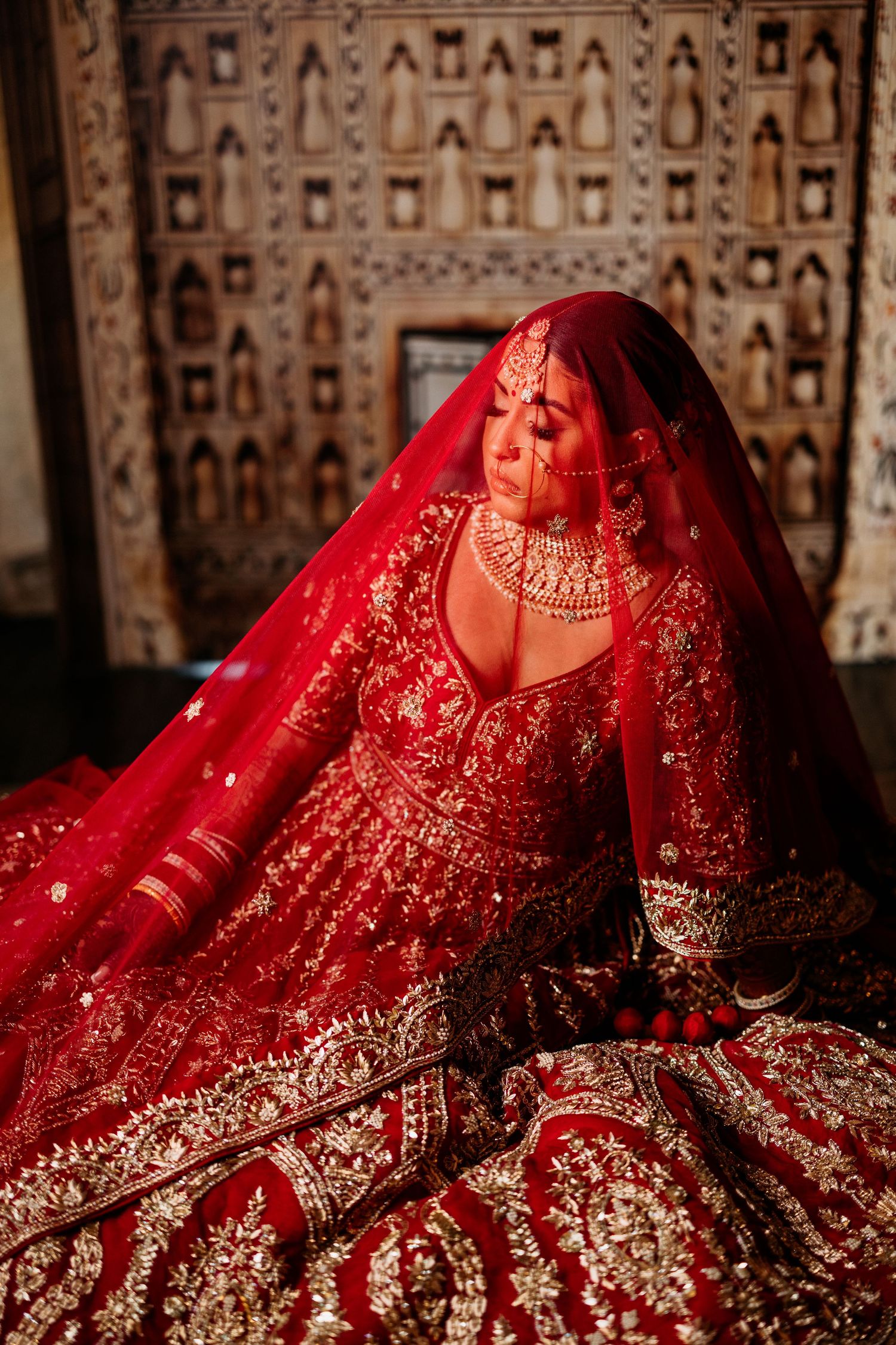 Traditional Indian bridal lehenga in deep red with intricate gold embroidery and embellishments displayed against patterned backdrop.