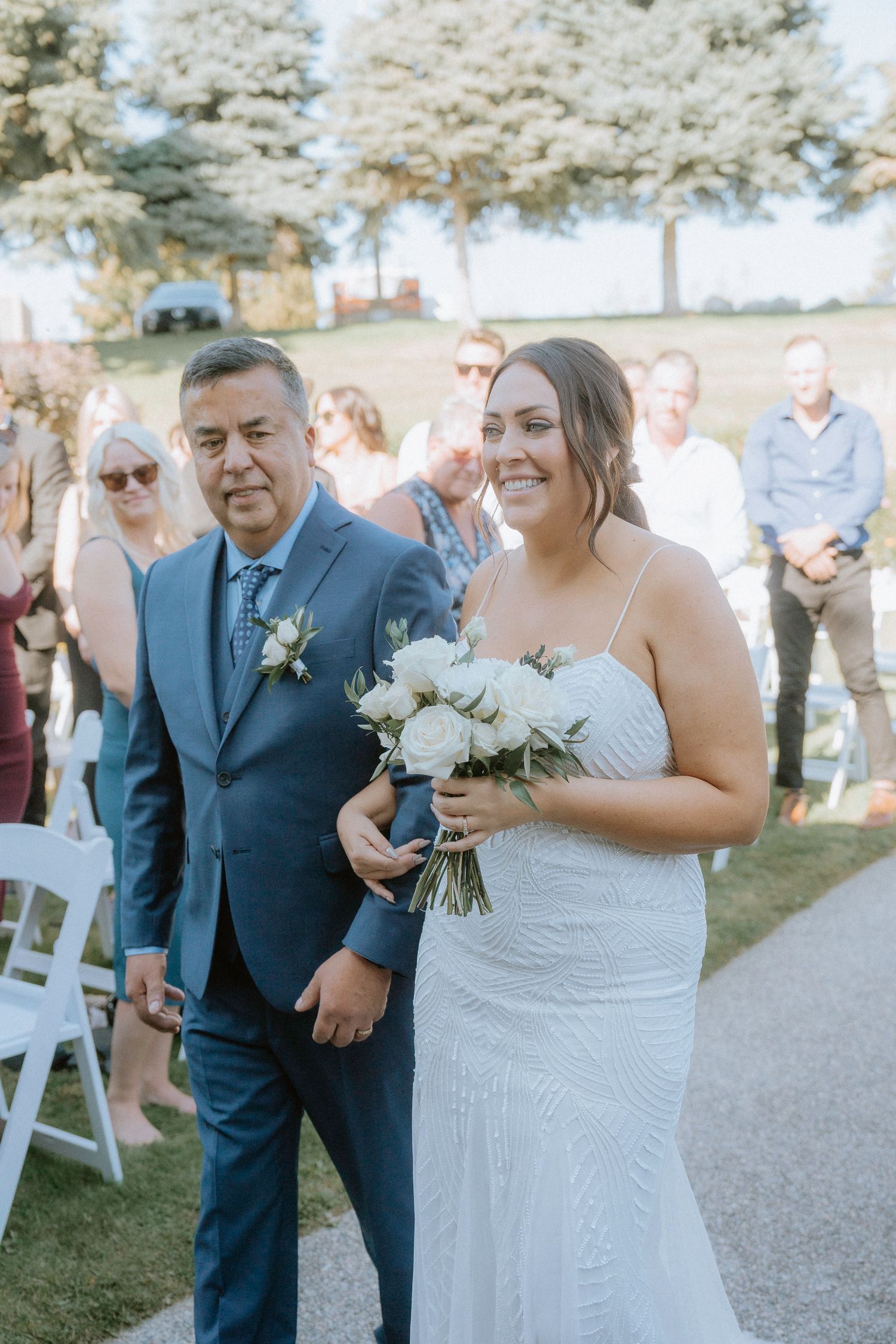 A wedding ceremony moment captured outdoors with white chairs and natural scenery in the background.