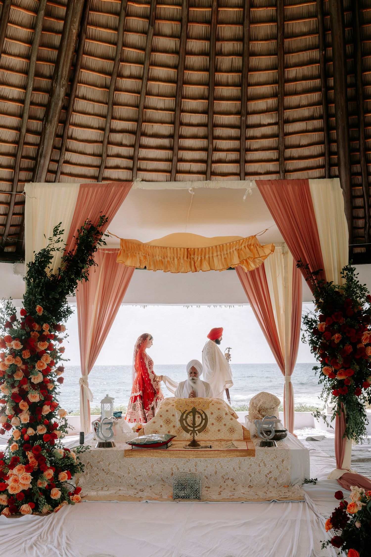 Elegant Indian wedding mandap decorated with peach drapes, floral arrangements and ornate white seating on a beach.