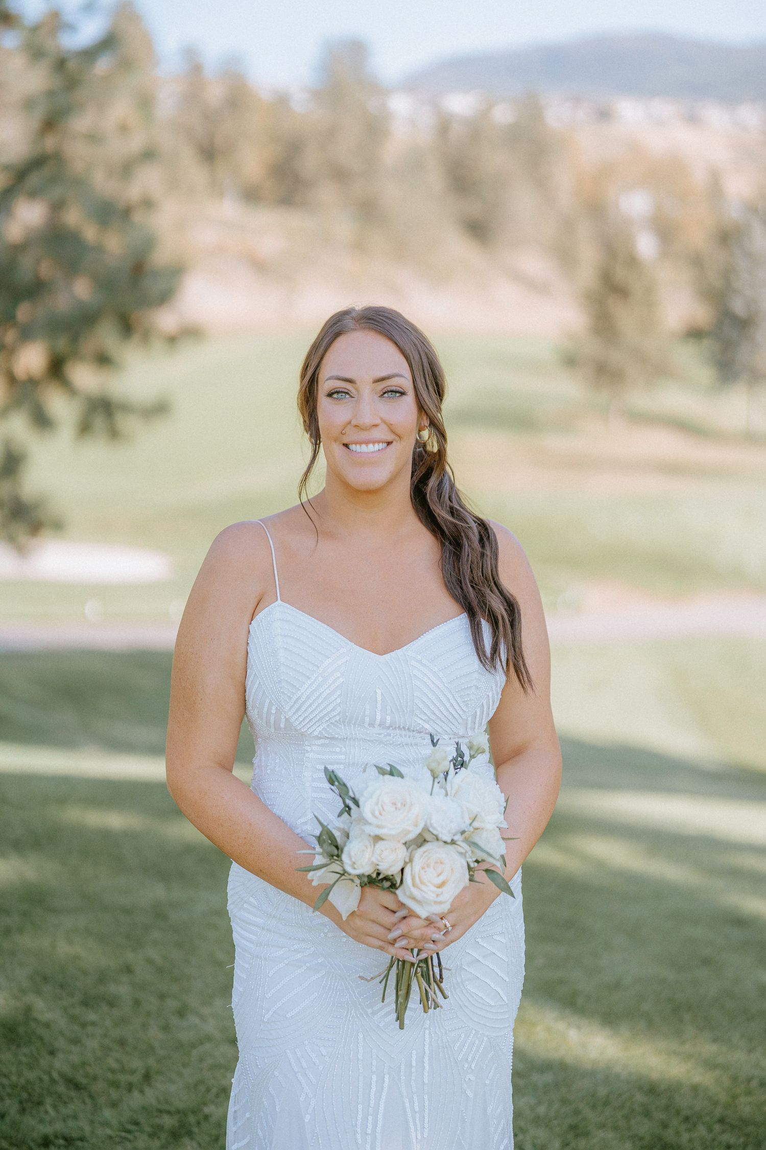 A bride in a white dress poses with a bouquet of white roses against a scenic outdoor backdrop.