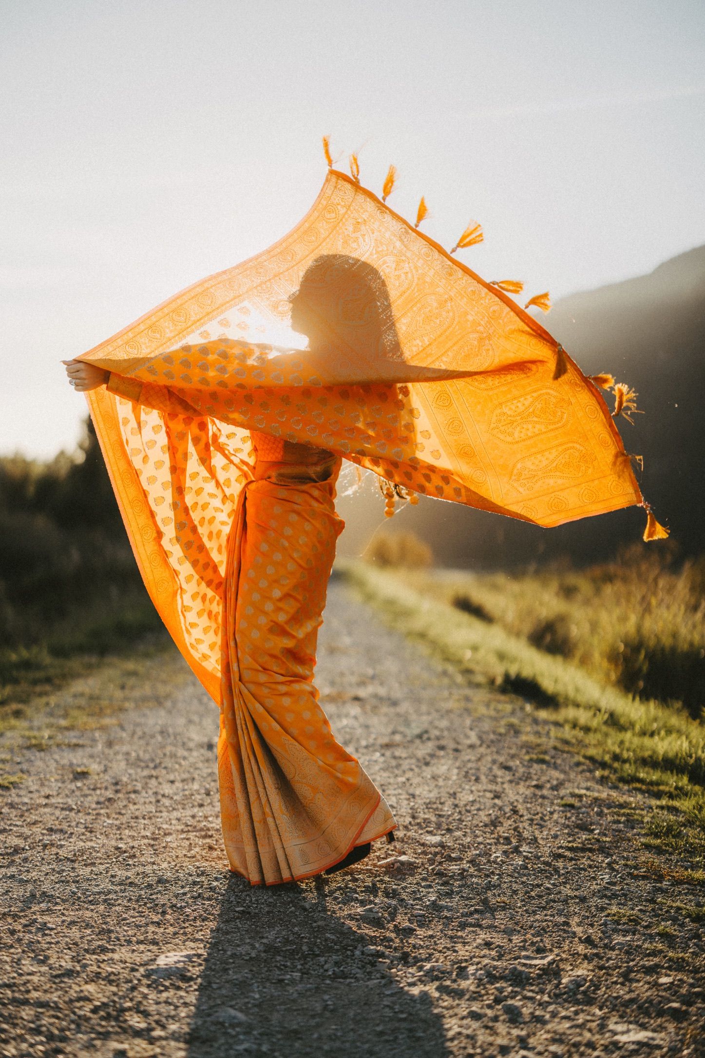 A flowing orange dress billows in the wind against a sunset backdrop on a dirt path.