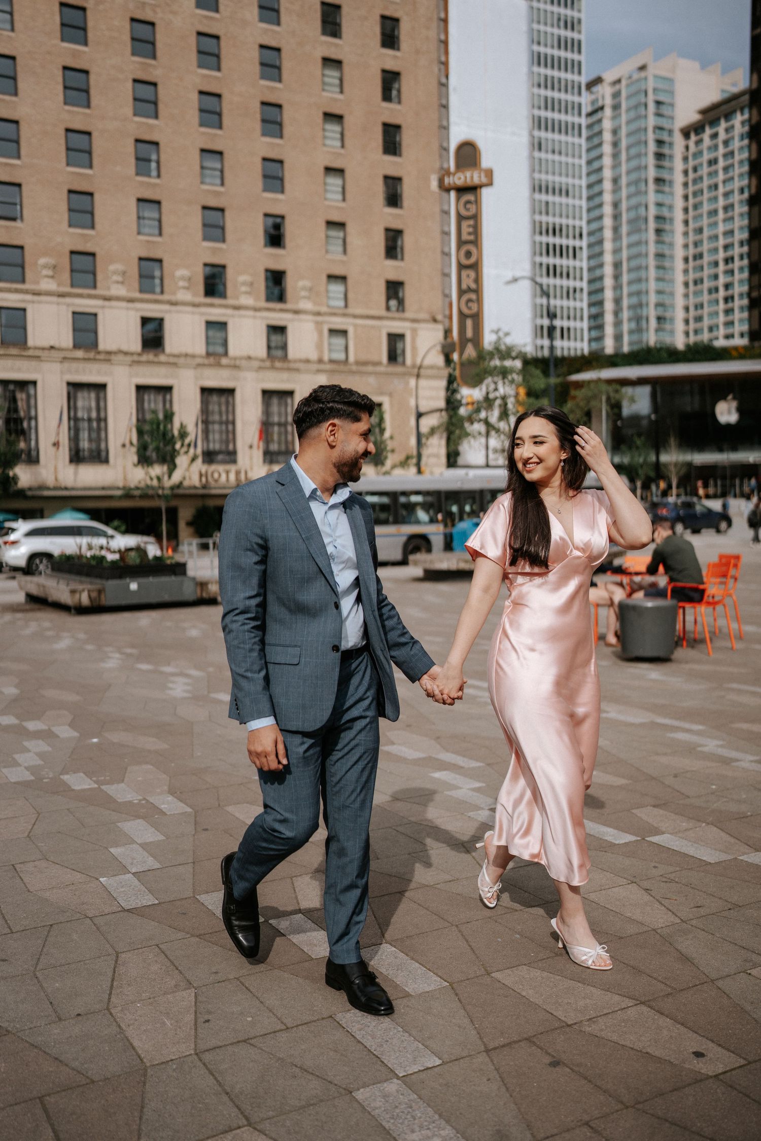 A couple in formal attire dances playfully on a city plaza with tall buildings in the background.