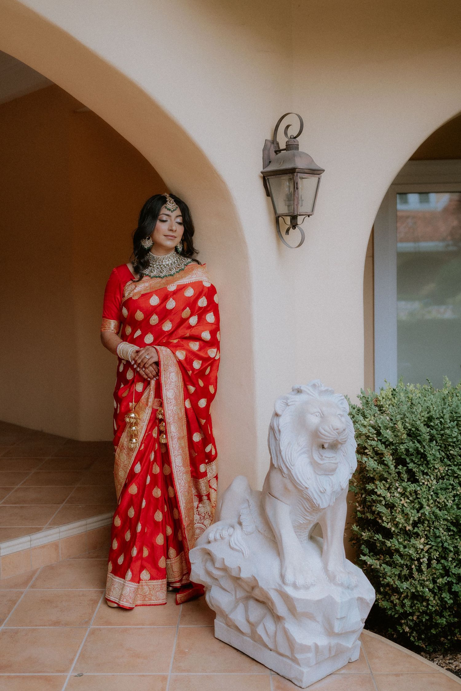 A person wearing a vibrant red and gold sari poses elegantly next to white stone lion statues in an outdoor setting.