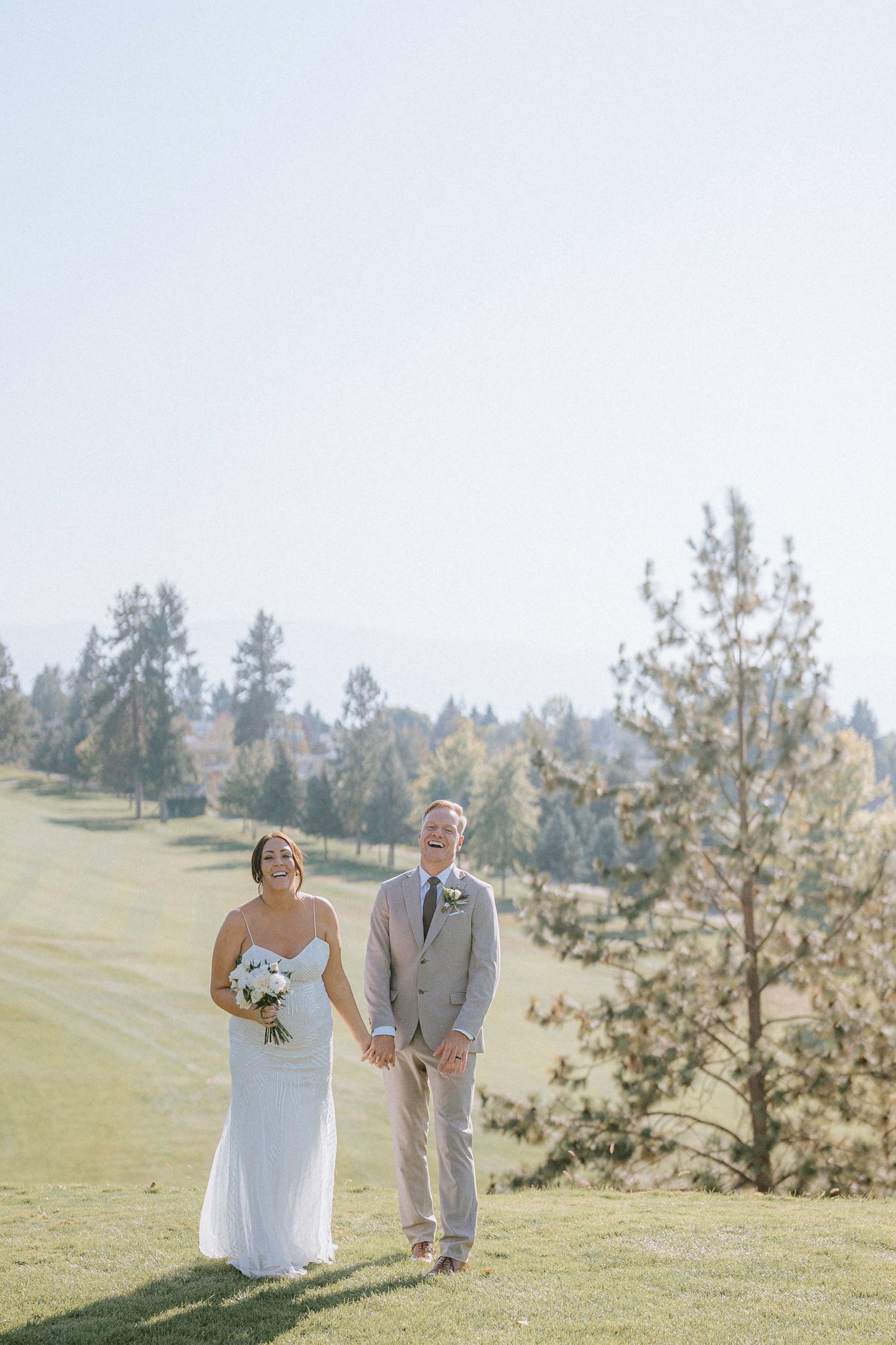 Wedding couple walking hand in hand across a sunlit grassy hill with mountain views in the background.