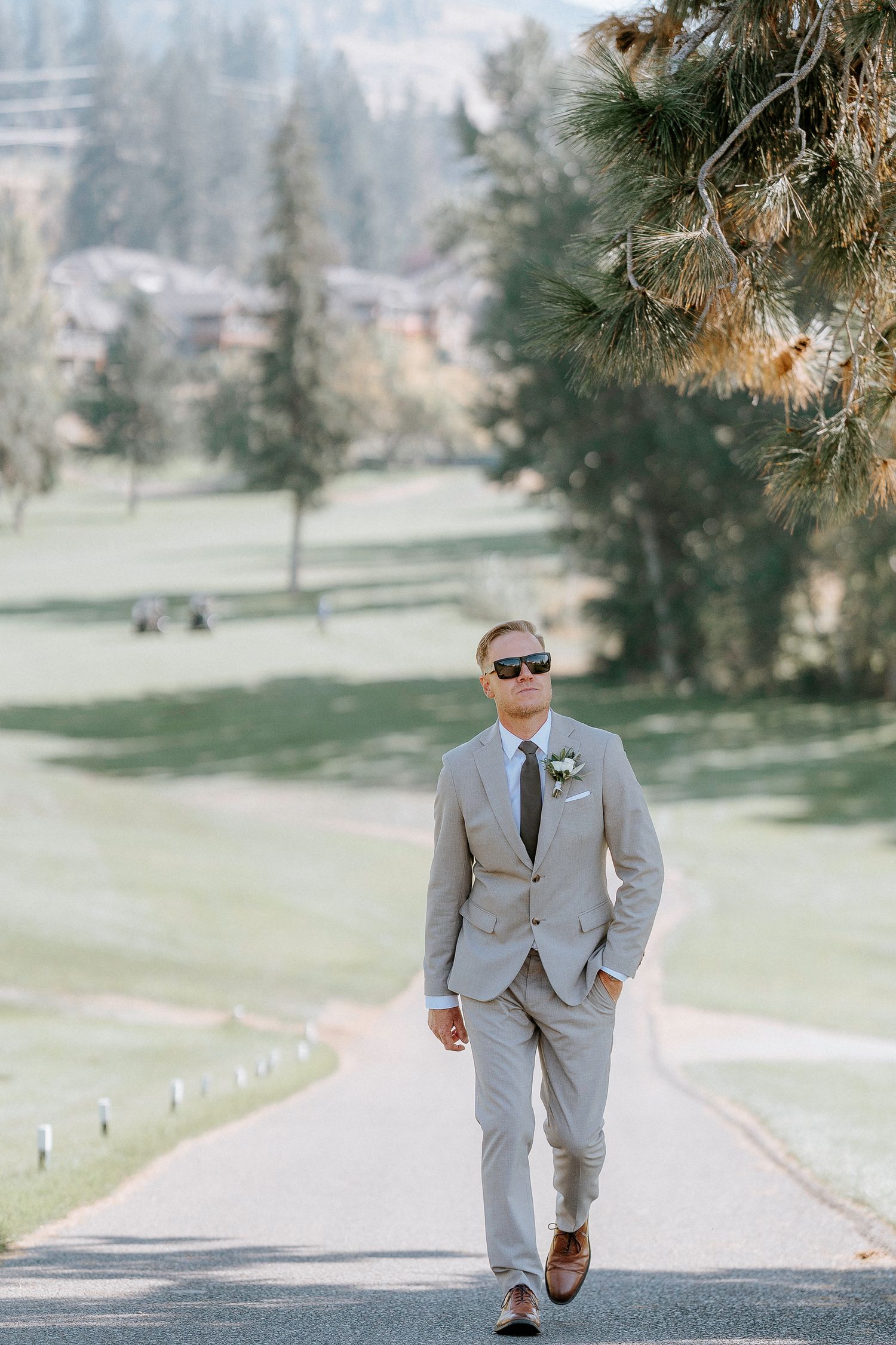 Groomsman in light gray suit and sunglasses walking confidently down a tree-lined path on a sunny day.