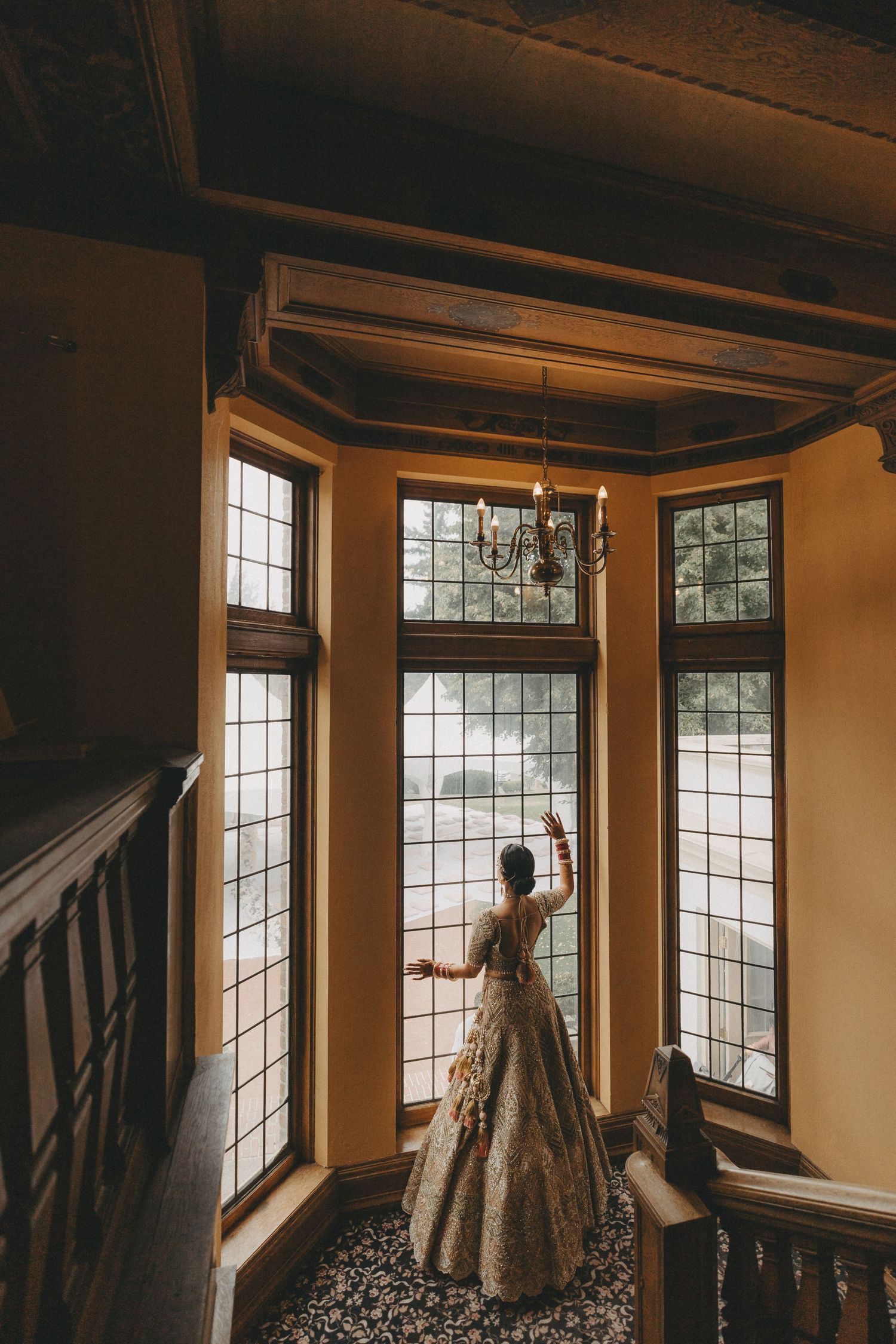 A person in an elegant ballgown gazes through ornate floor-to-ceiling windows in a vintage mansion's bay window alcove.