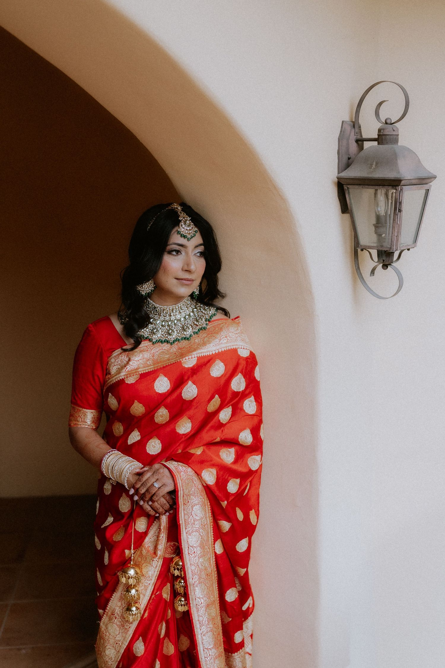 A person wearing a vibrant red and white polka dot saree with gold embellishments stands against a neutral wall with sconces.