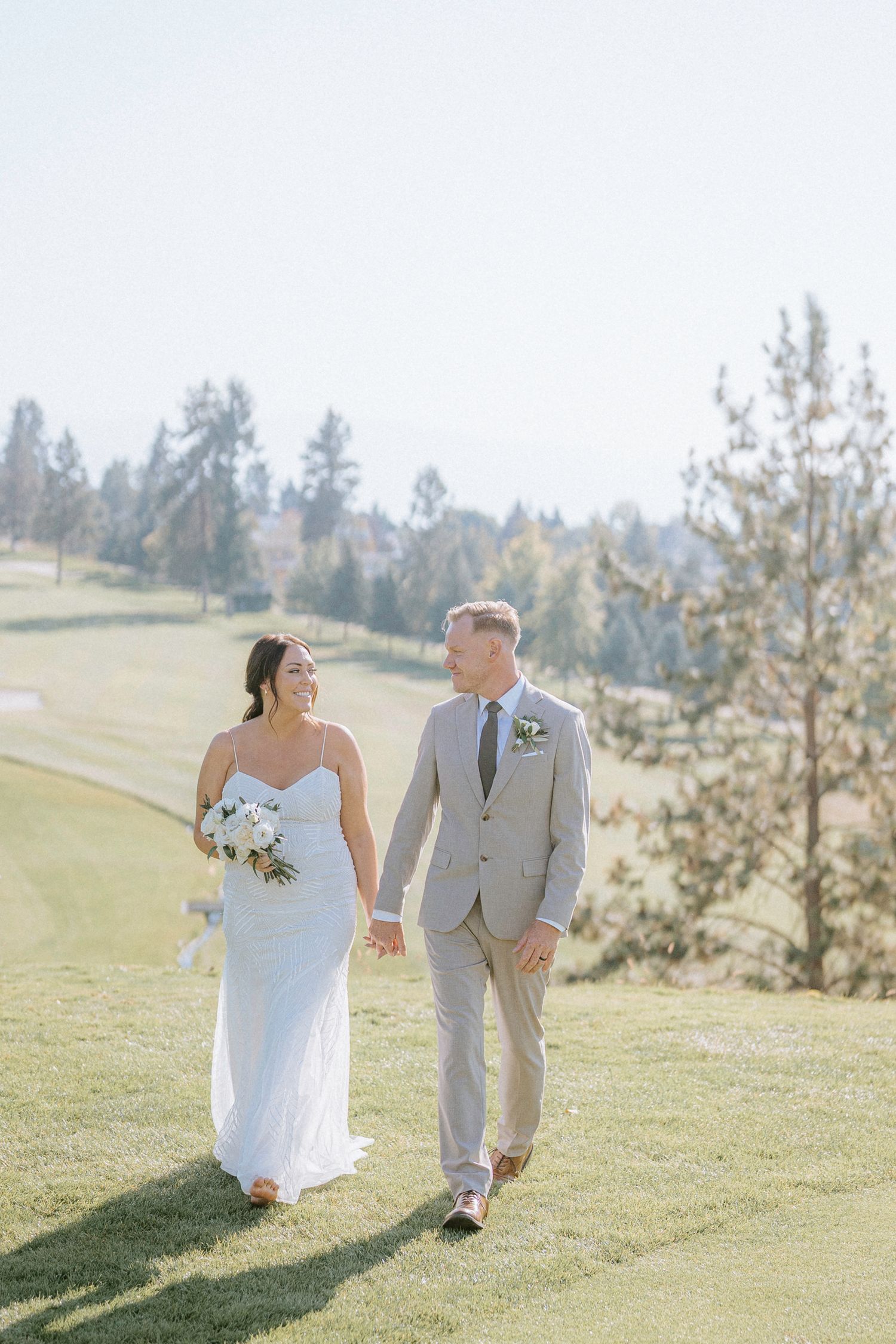 A couple in wedding attire walks together on a sunlit grassy hillside during their outdoor wedding celebration.