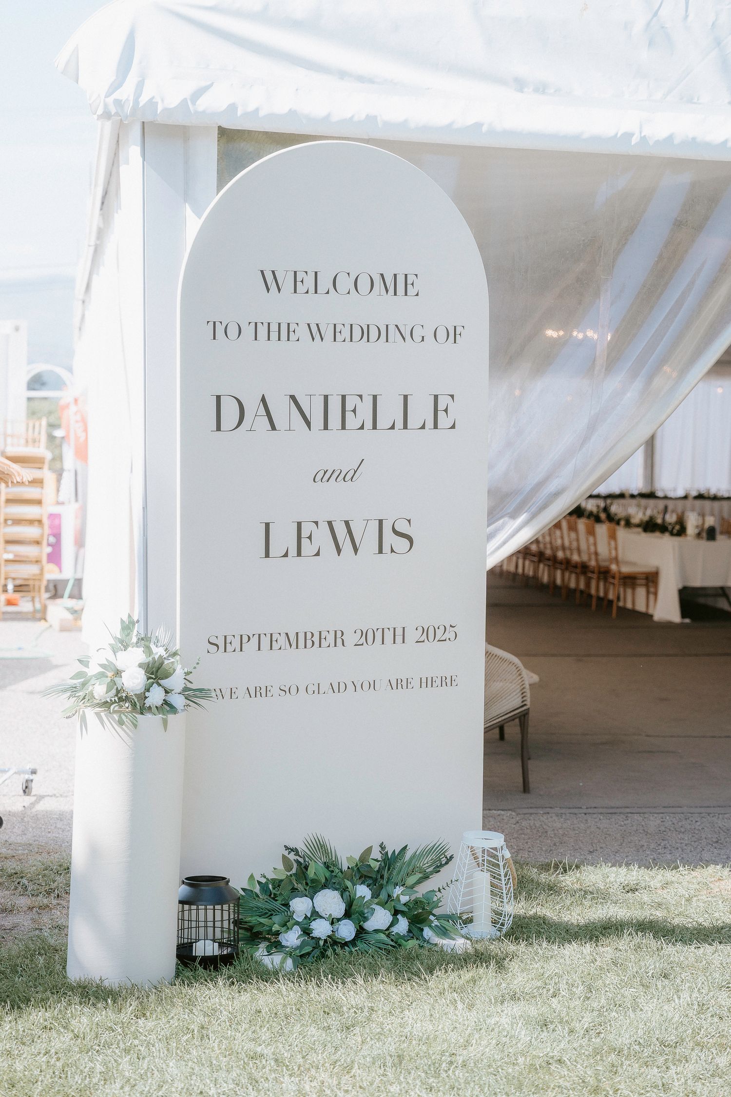 Elegant white wedding welcome sign with floral arrangements displaying names Danielle and Lewis for September 2025 ceremony.