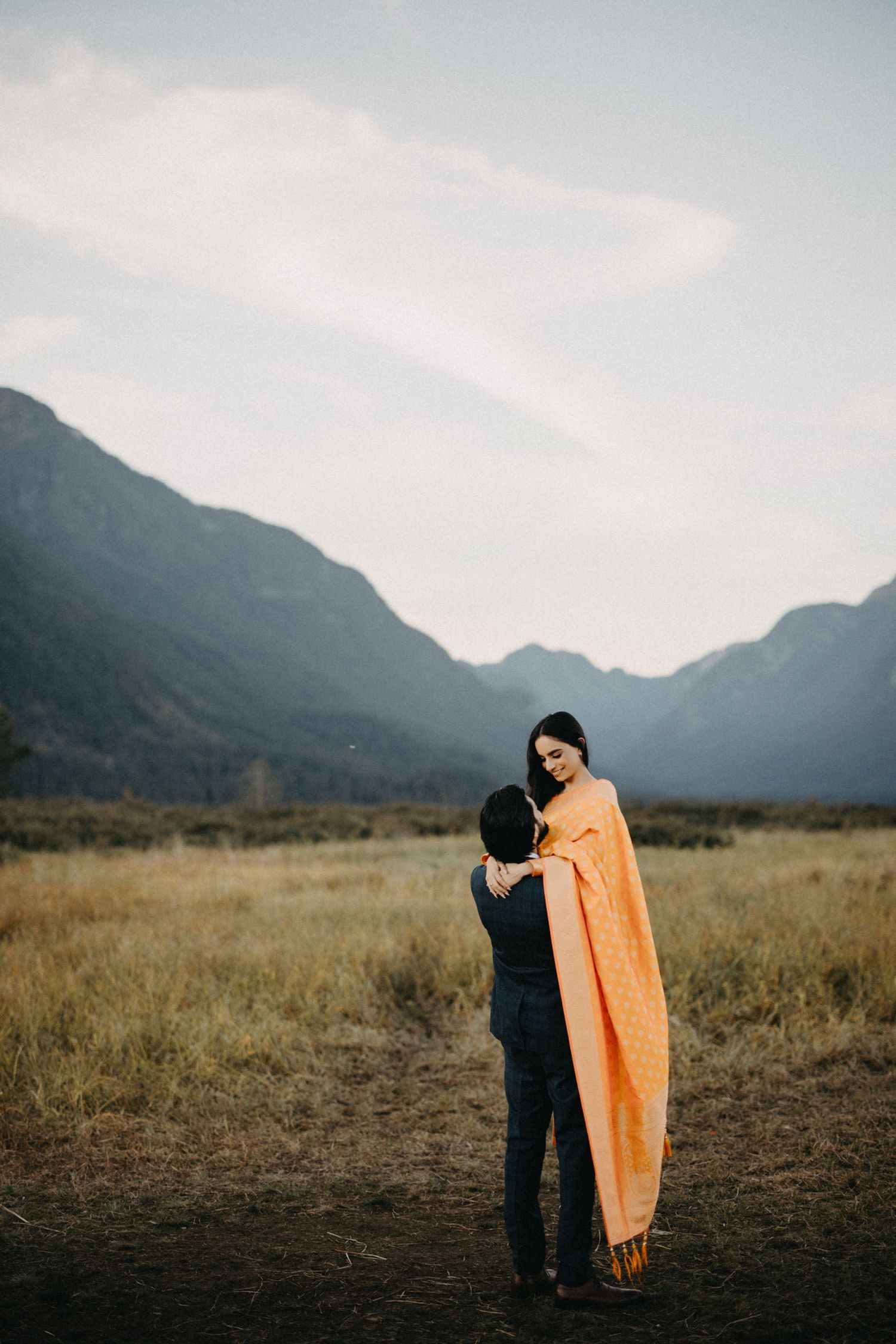 Two people wrapped in an orange blanket stand together in a misty mountain field at dusk.