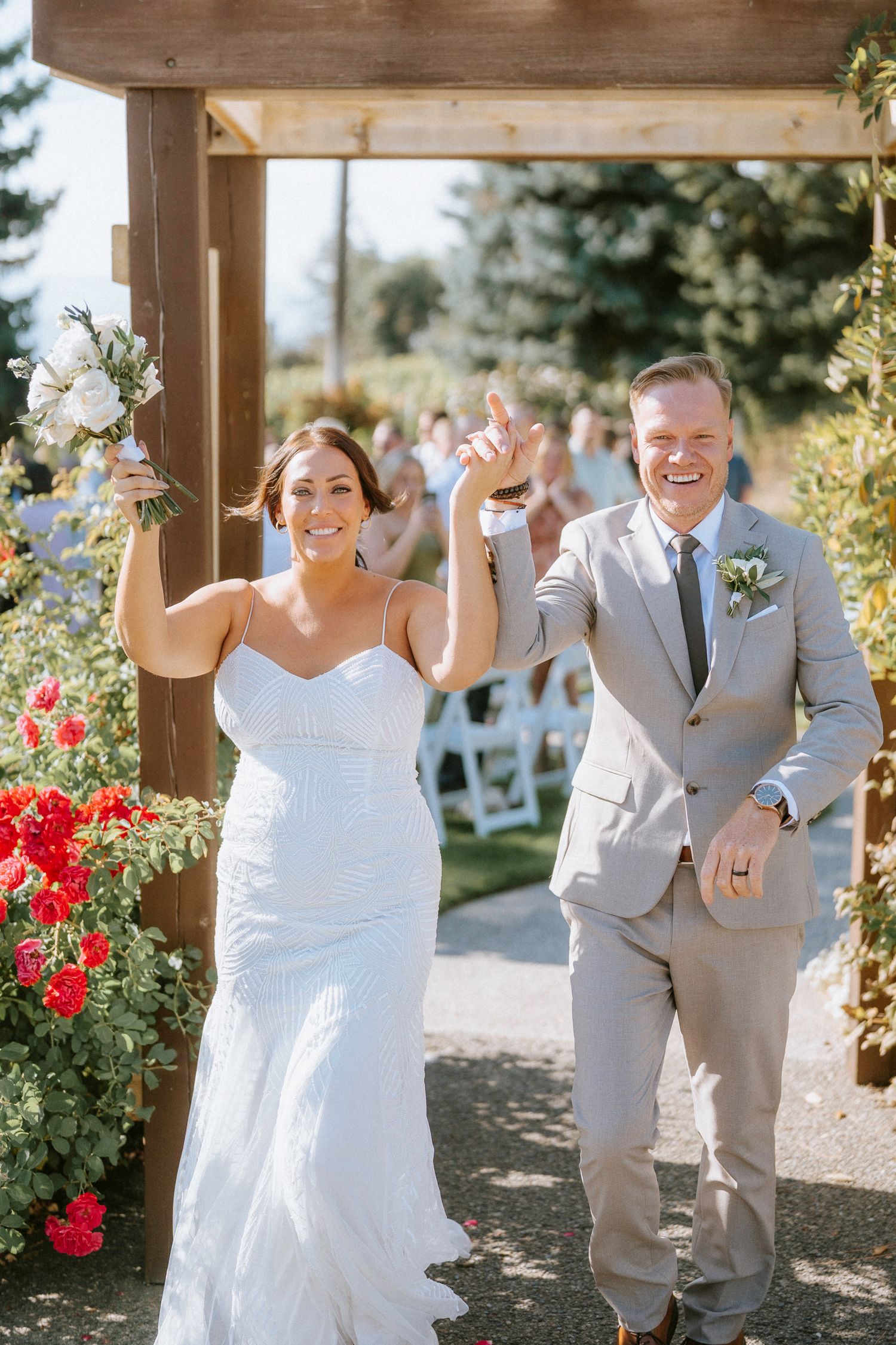 Newlywed couple celebrates joyfully while walking down outdoor garden path with red roses and wooden pergola.