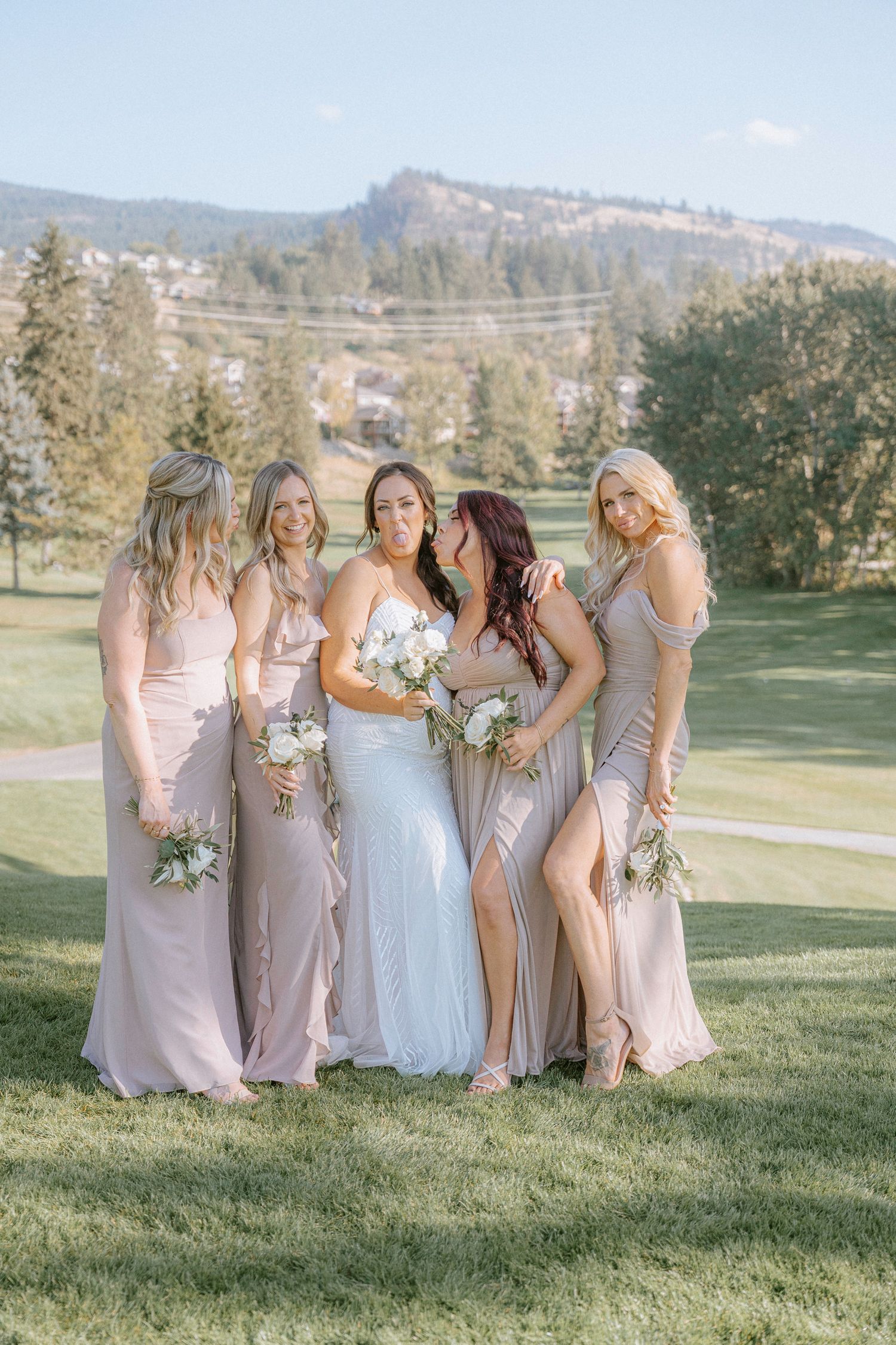 Bridesmaids in blush pink dresses pose with bouquets on a hillside with mountain views.