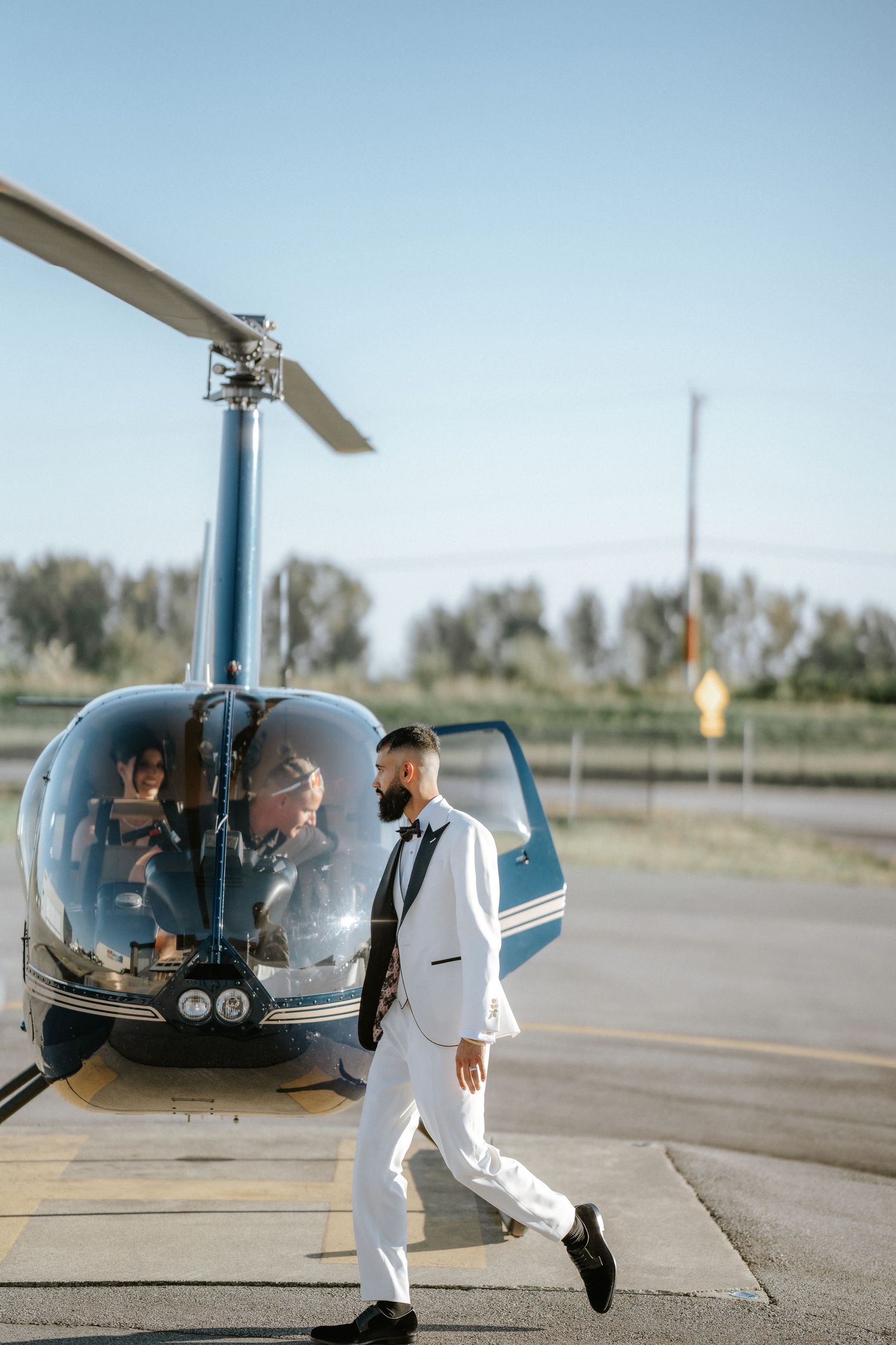 A couple in white outfits walking towards and boarding a small black helicopter on an airfield tarmac.