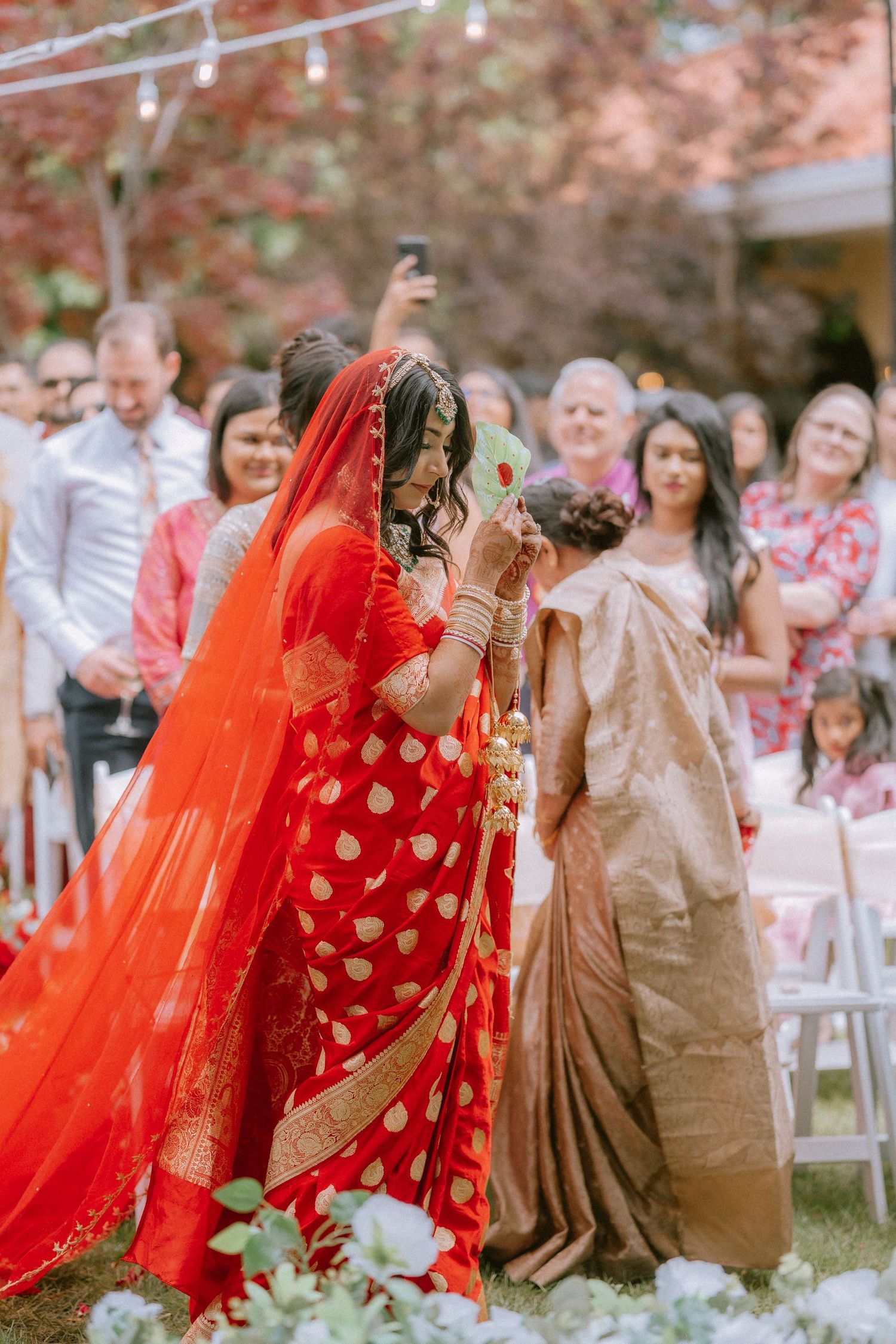 Wedding guests in colorful saris and traditional Indian attire gather outdoors under cherry blossoms.