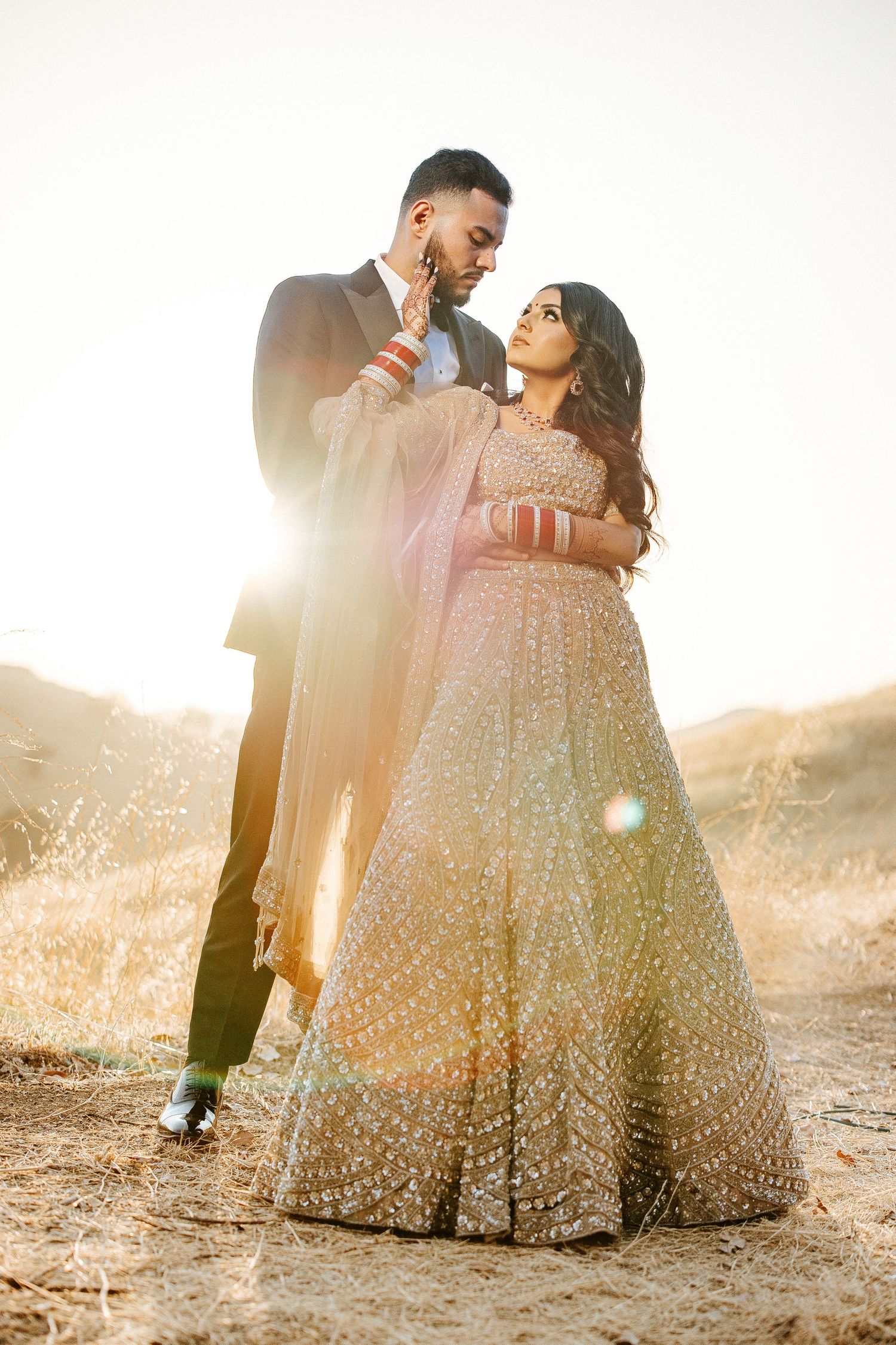 A couple in traditional Indian wedding attire embraces during a golden hour photoshoot in an outdoor setting.