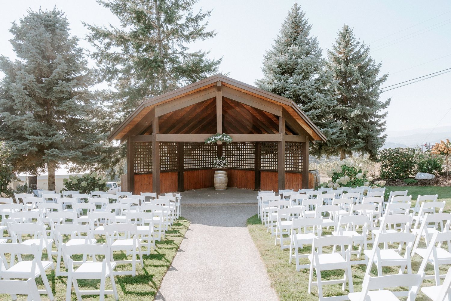 Rows of white folding chairs arranged outdoors for a wedding ceremony with evergreen trees and a wooden gazebo in the background.