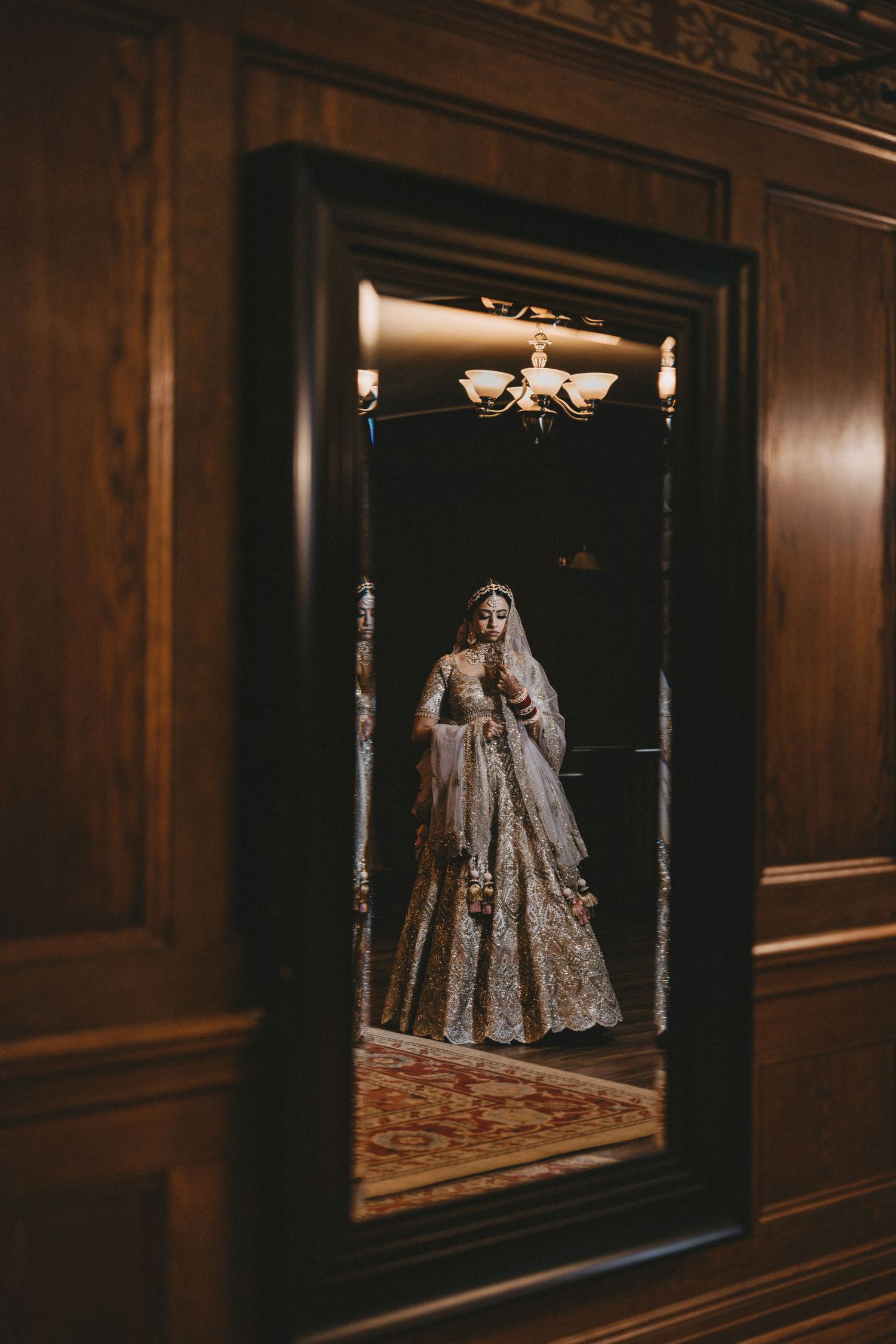 Reflection in an ornate wooden mirror shows an elegant figure in a detailed gold lehenga dress in a dimly lit room.