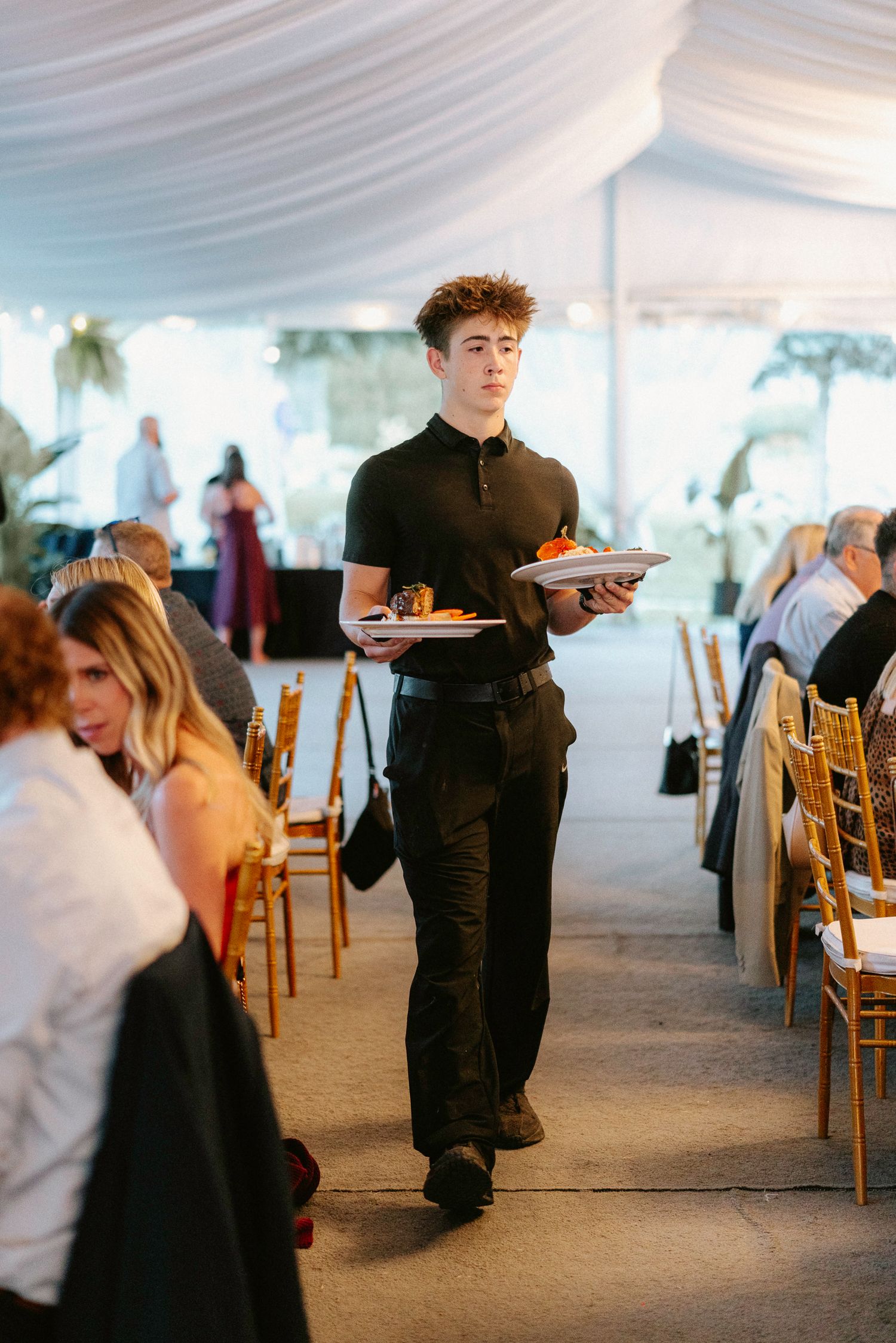 Wedding reception staff in black attire serve guests in an elegant white tent setting.