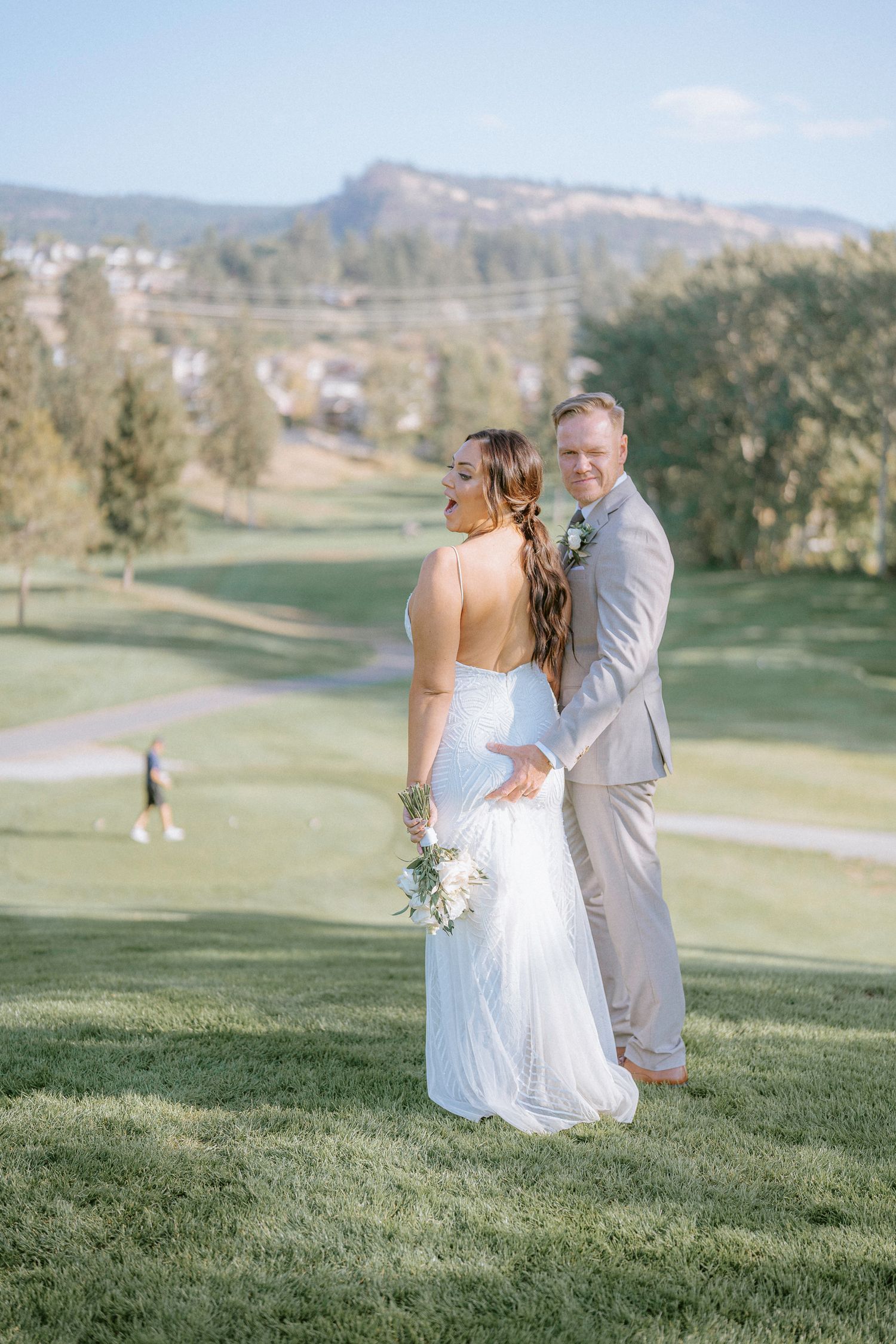 Couple in wedding attire embracing on a scenic golf course with mountains in background.