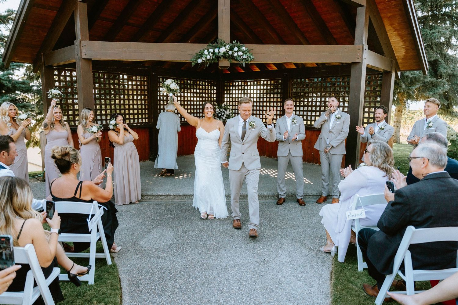 Wedding guests gather under a wooden pavilion with white chairs set up for the ceremony.