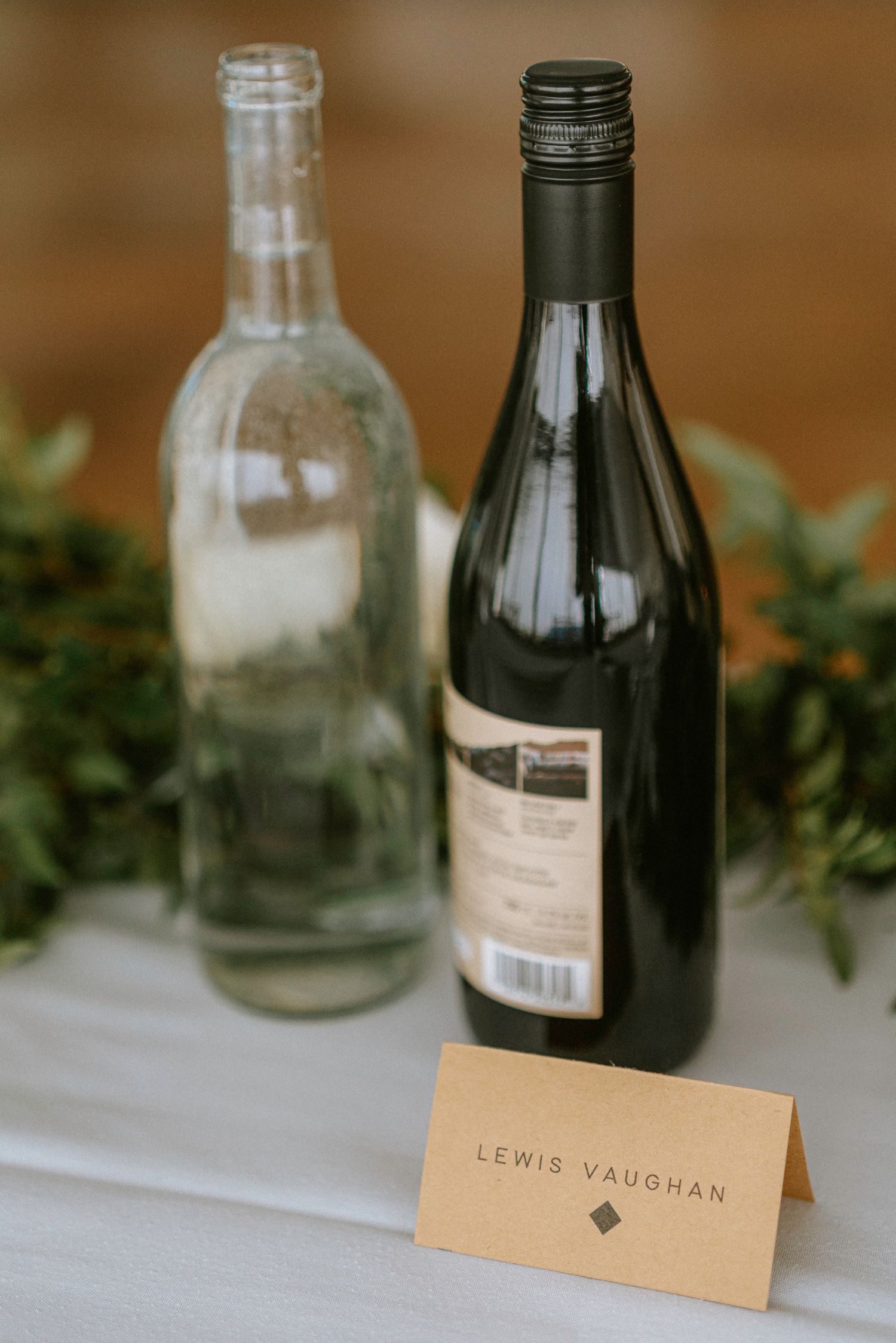 Wedding reception place cards and wine bottles arranged on wooden table with greenery accents.