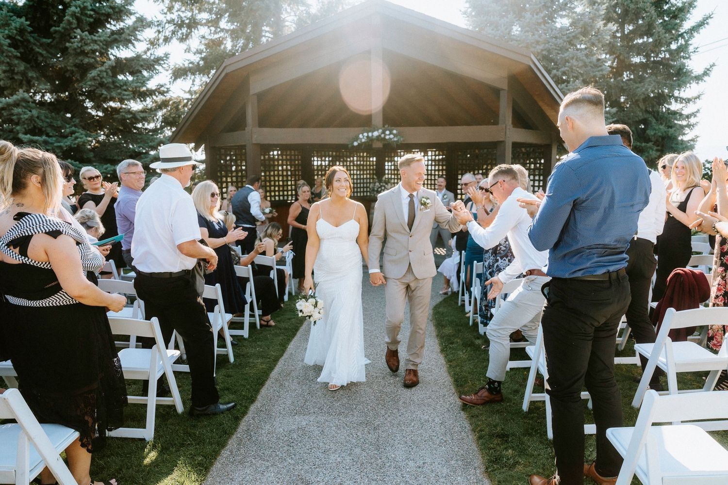 Wedding ceremony procession walking down outdoor aisle with gazebo and evergreen trees in background.