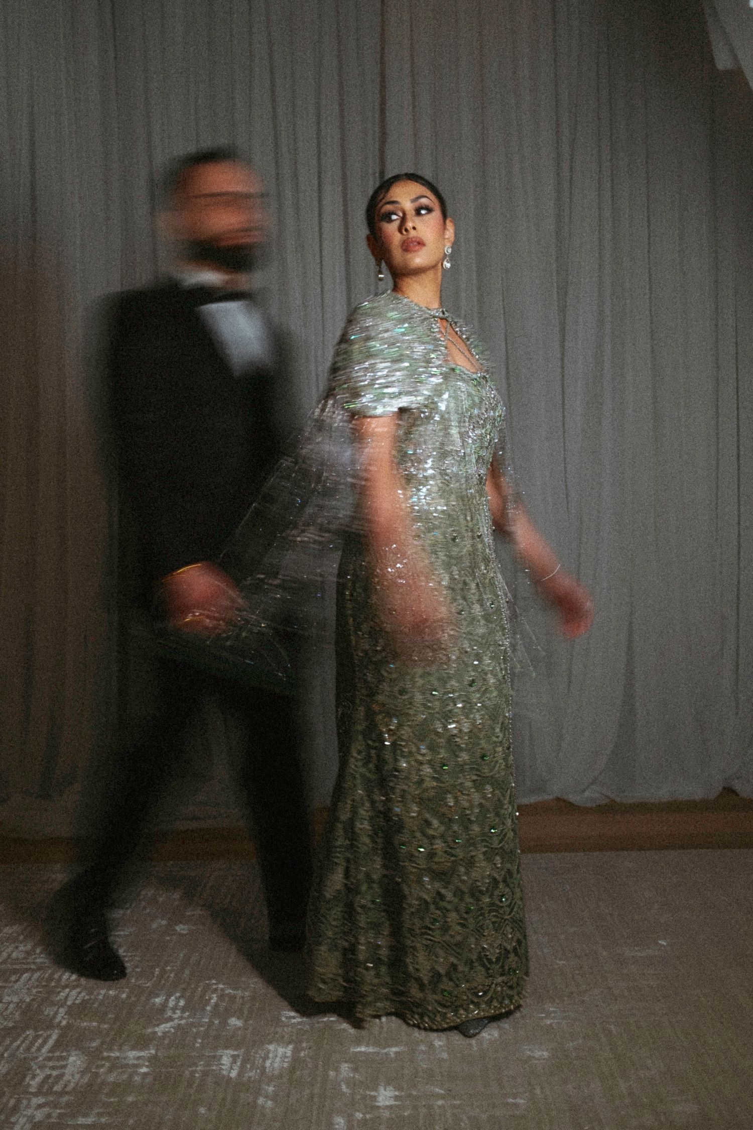 Motion-blurred figures dance in a room with curtains, one wearing a sparkly silver dress while others move around them.