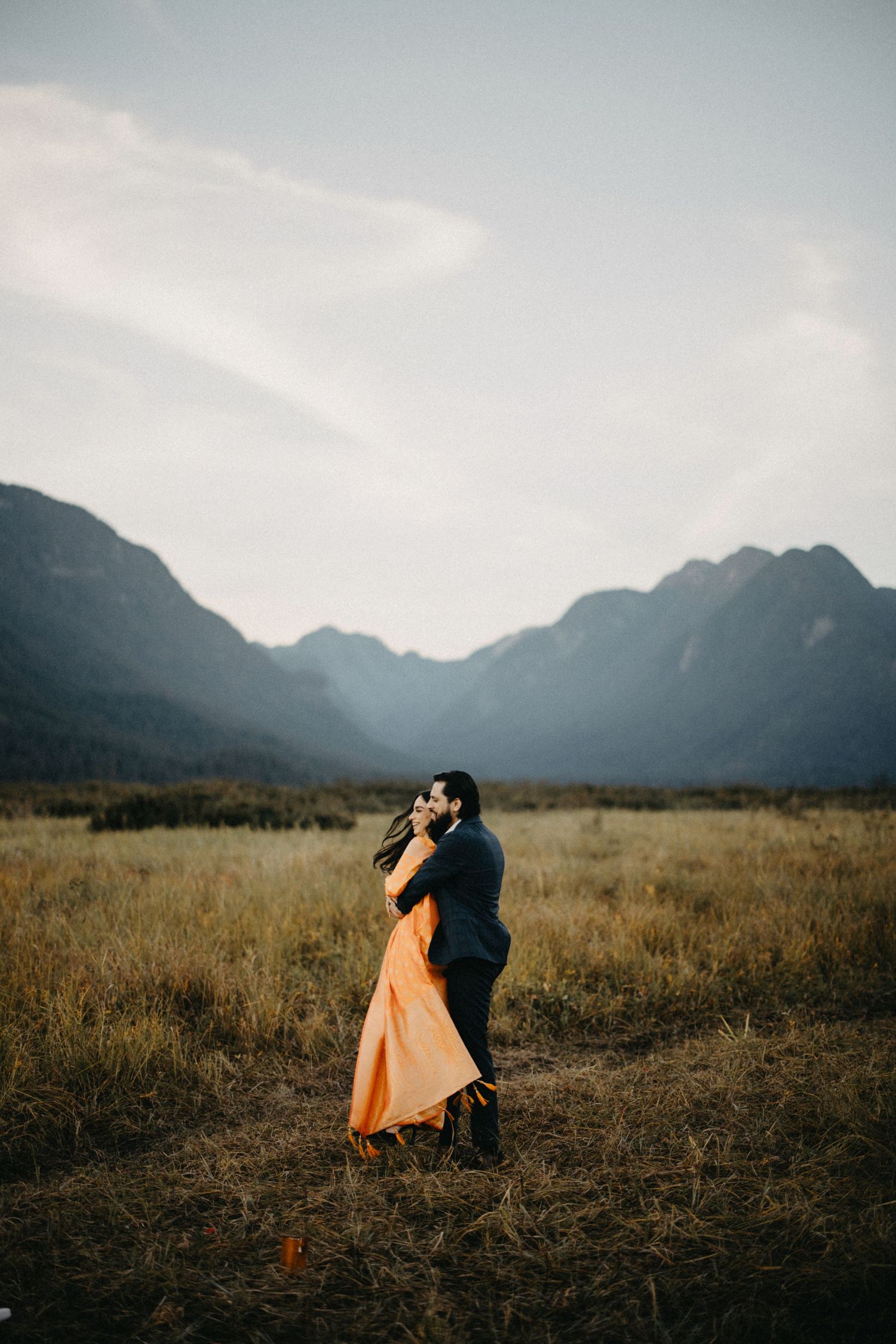 A couple embraces in silhouette against a dramatic mountain backdrop during golden hour in a grassy meadow.