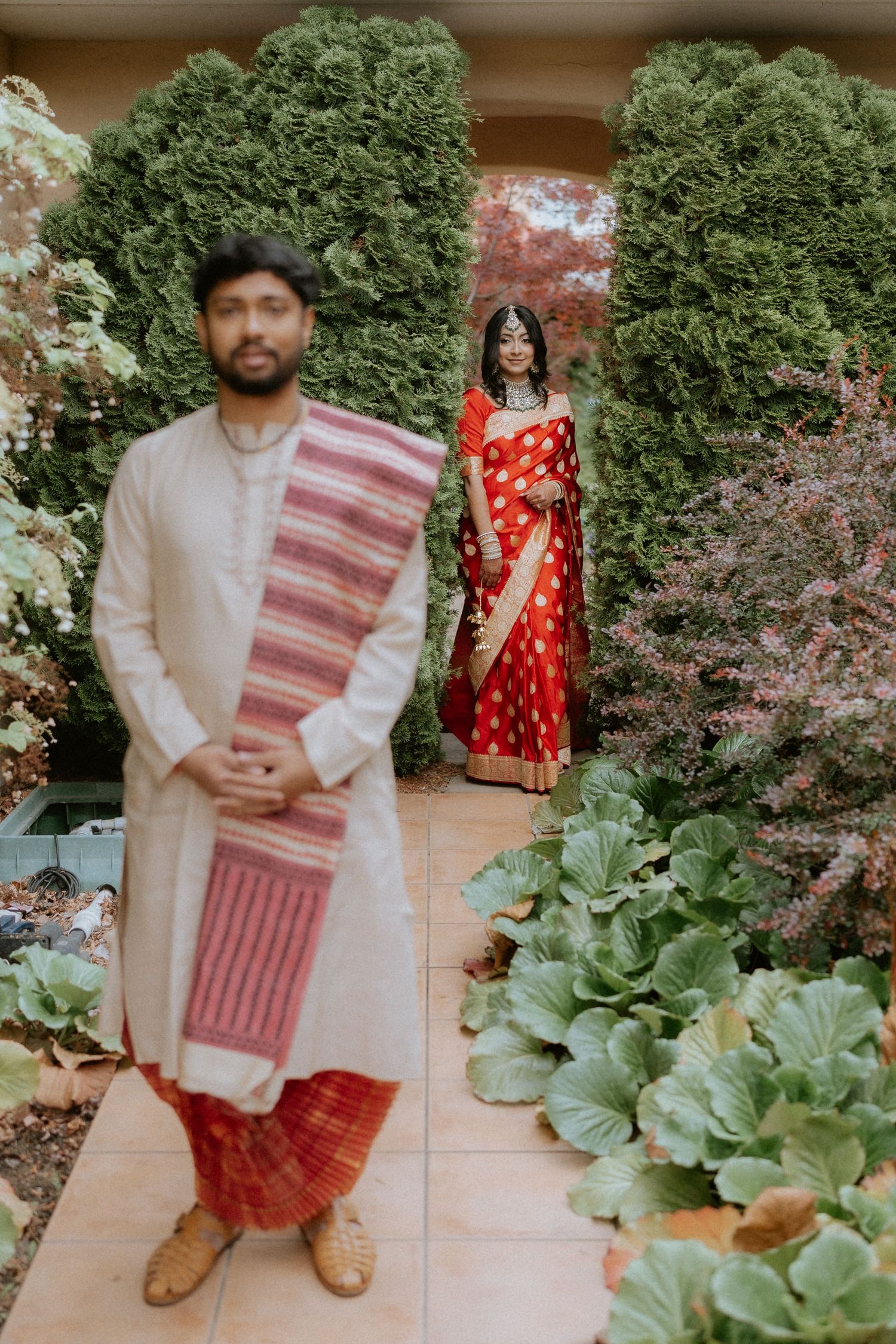 A couple poses in traditional Indian attire against lush greenery and garden plants in a series of romantic photos.