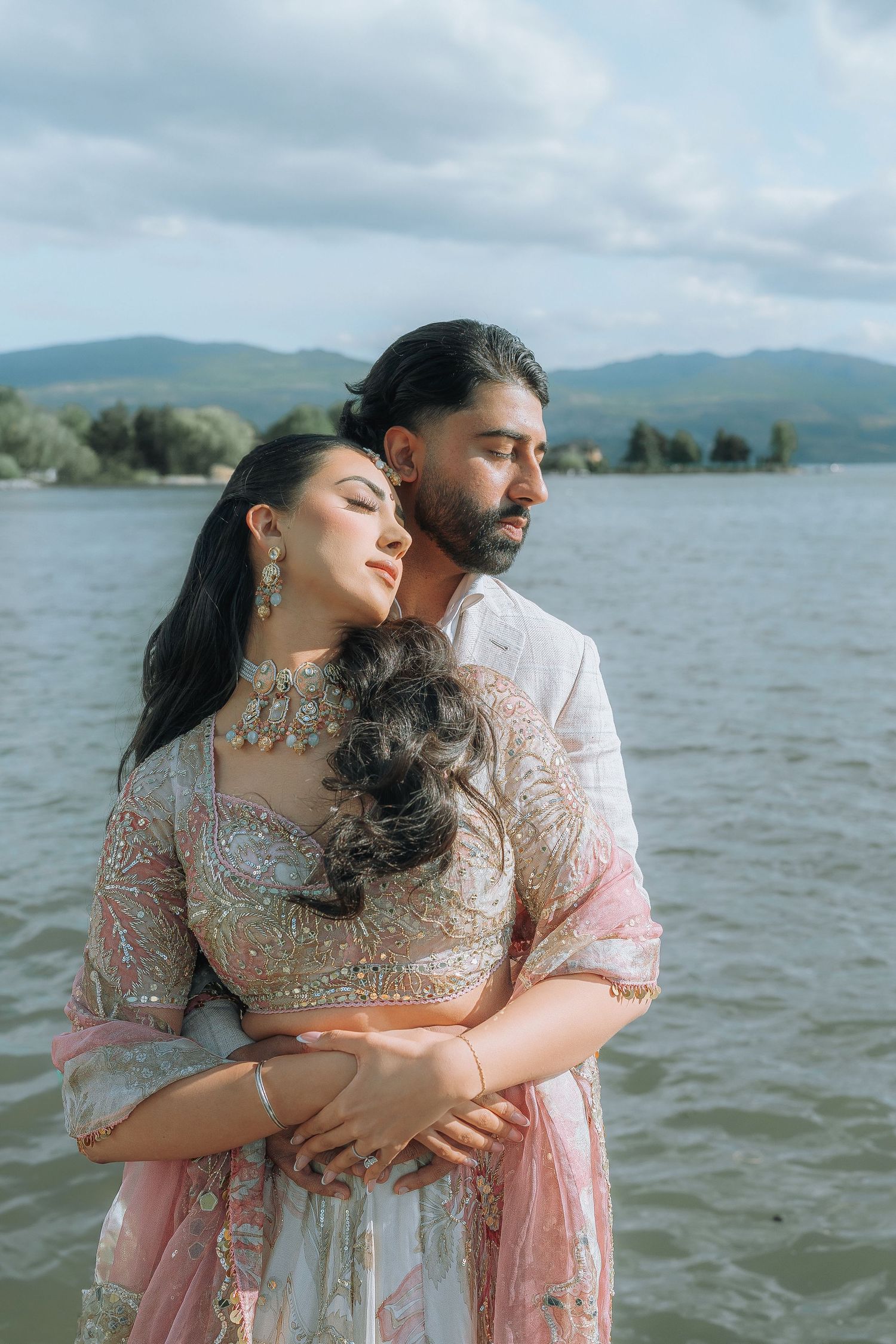 Couple in coordinated pink outfits embrace lovingly near a peaceful lake with mountains in the background.