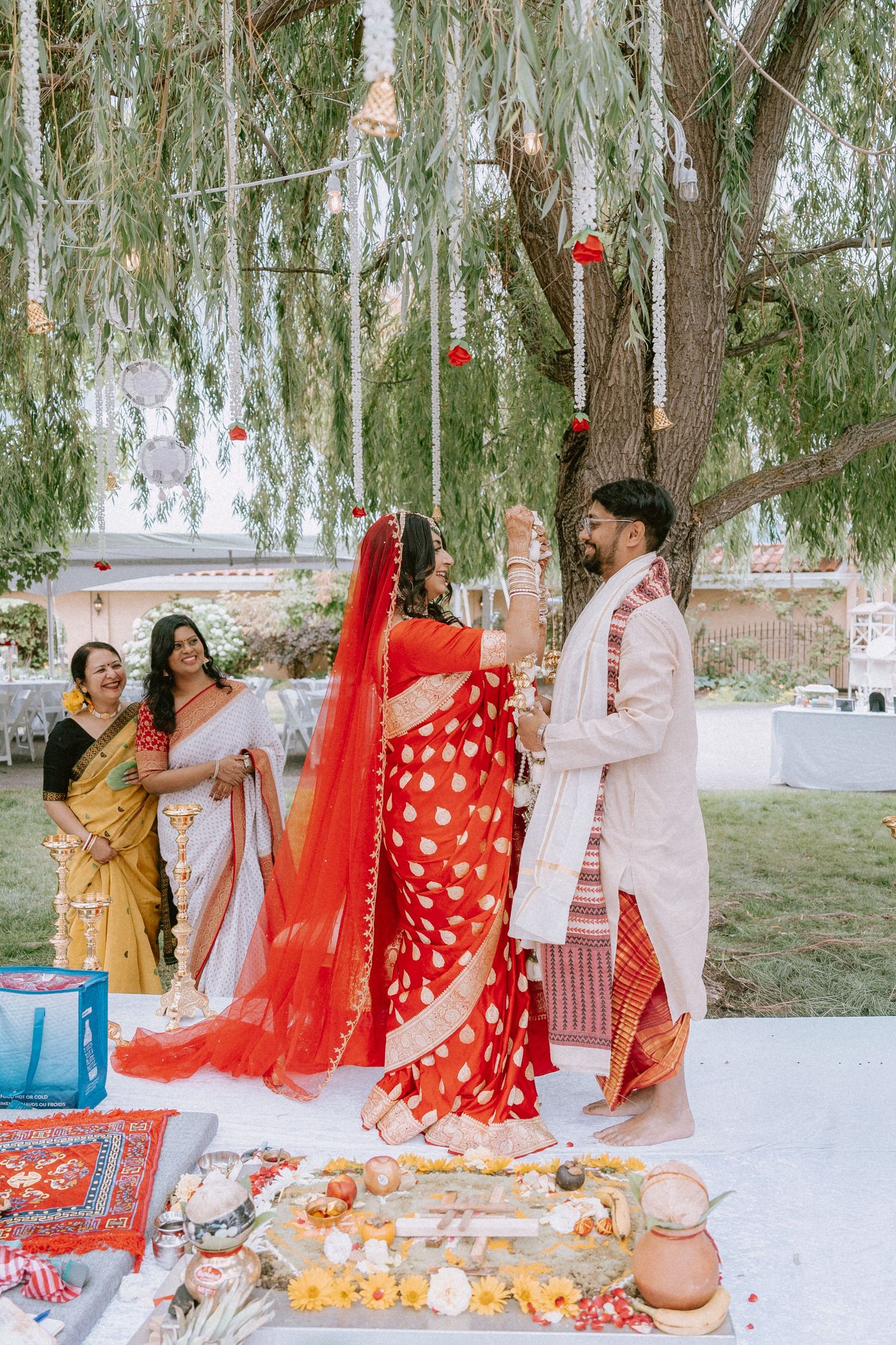 Wedding celebration with guests in traditional Indian attire gathered under willow trees.
