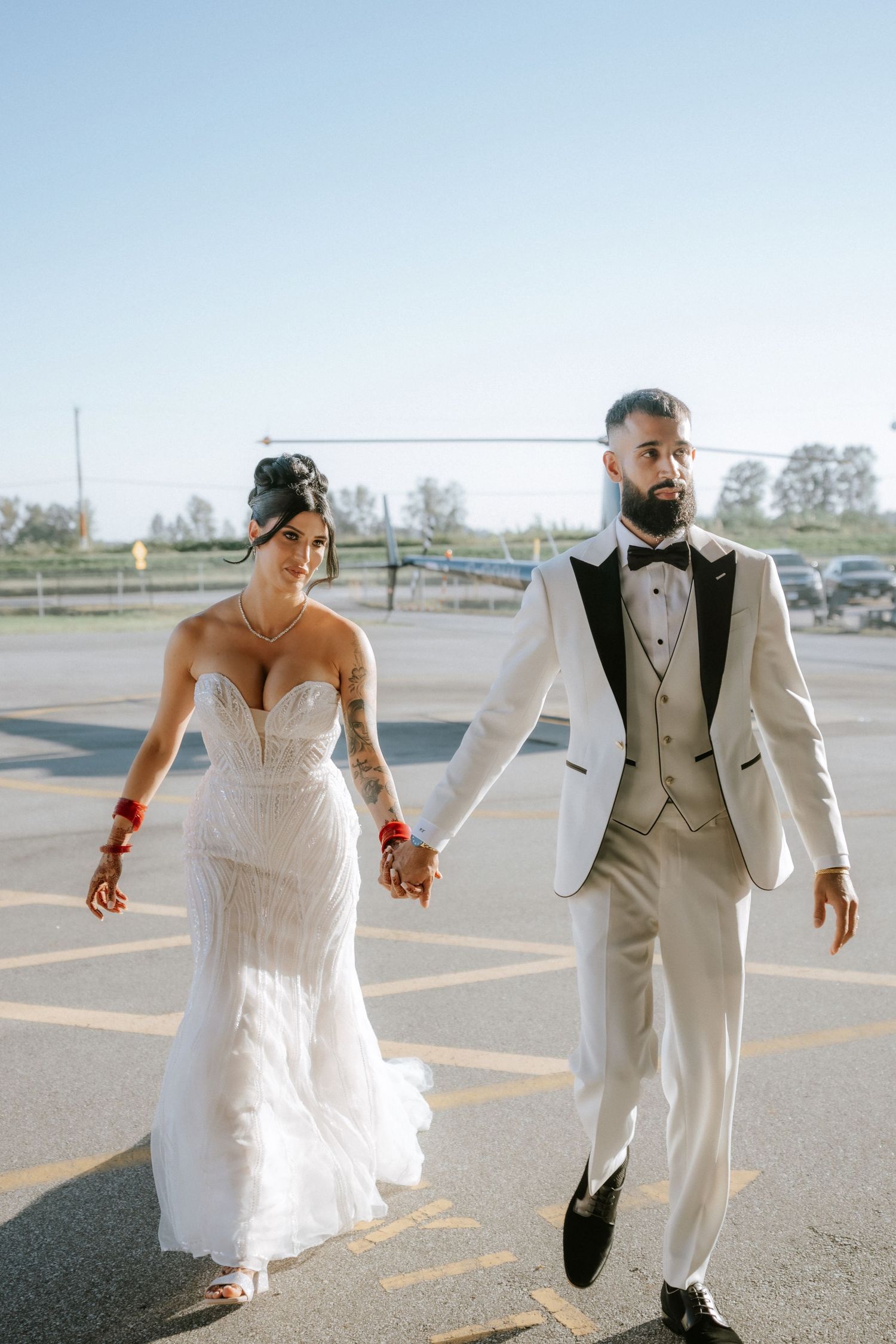 A bride and groom walk playfully together in a parking lot wearing white and gray formal wedding attire.