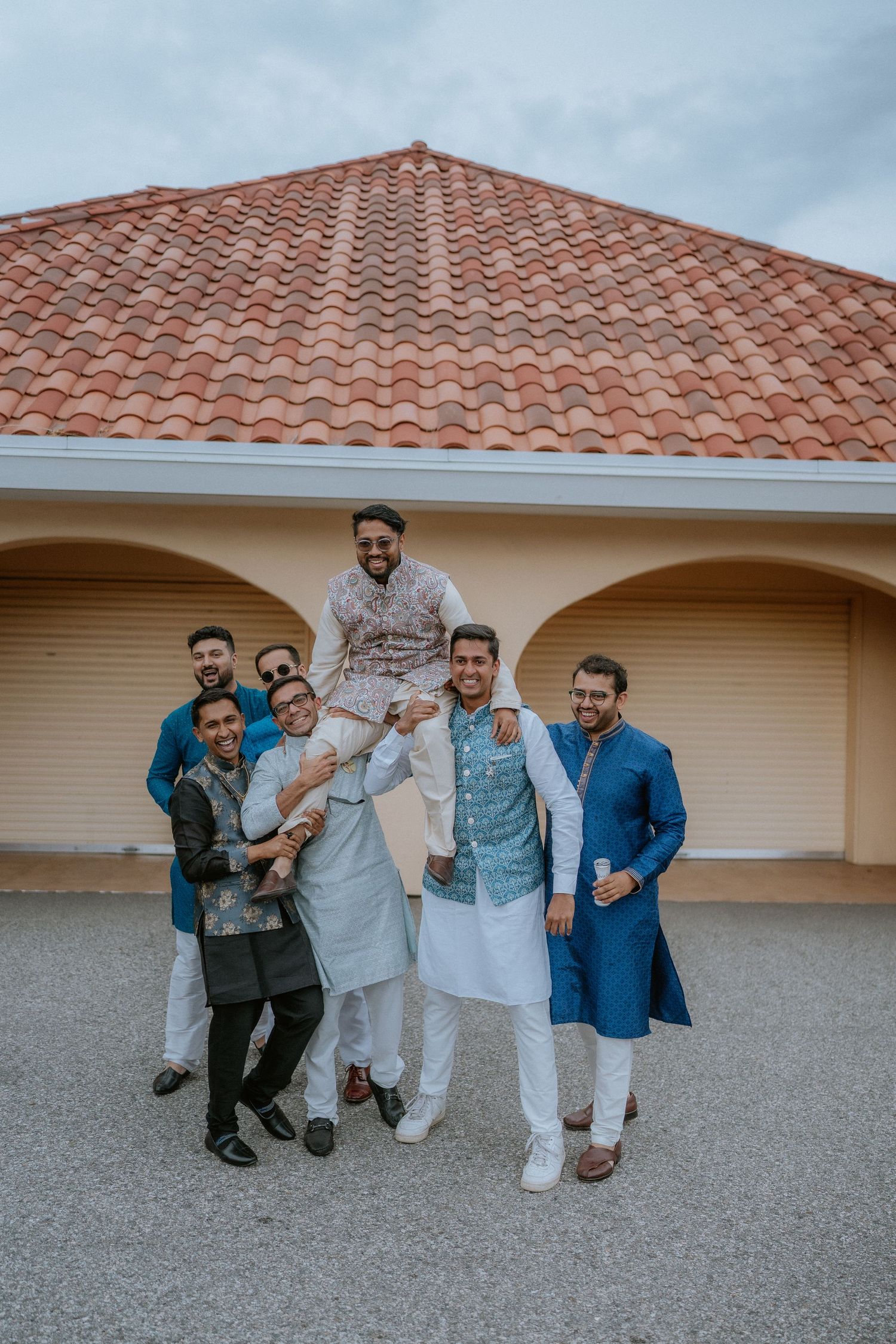 A group poses playfully together against a beige wall with a terracotta tiled roof in the background.