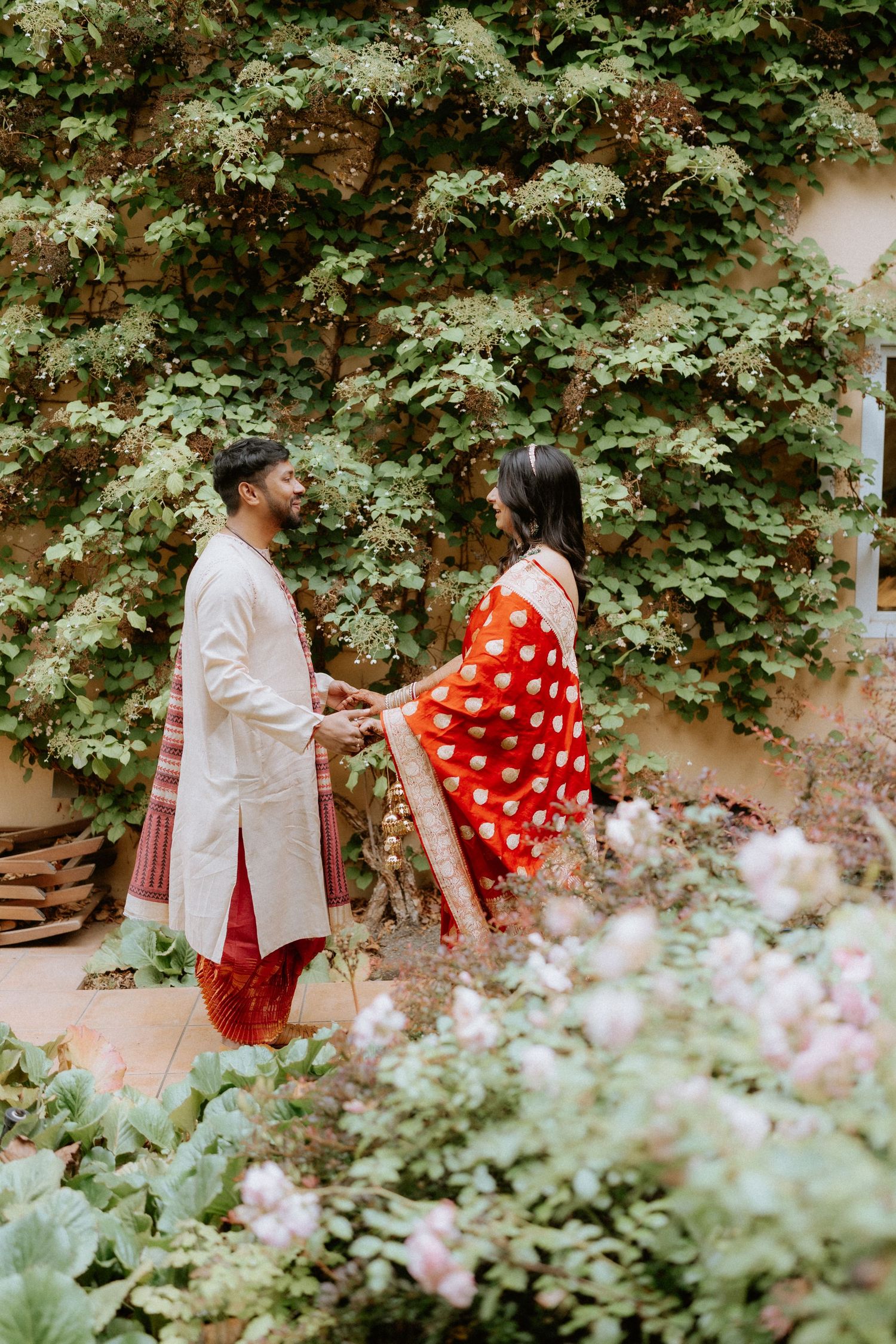 A couple shares a romantic moment in a garden setting surrounded by lush greenery and pink flowers.