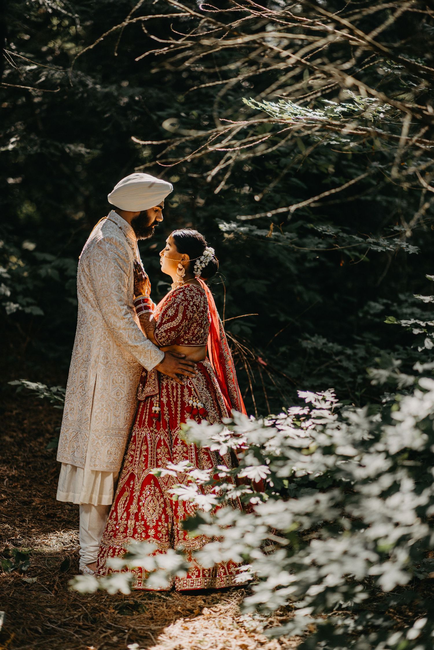 A couple in traditional Indian wedding attire stands together in a forest setting surrounded by delicate white flowers.
