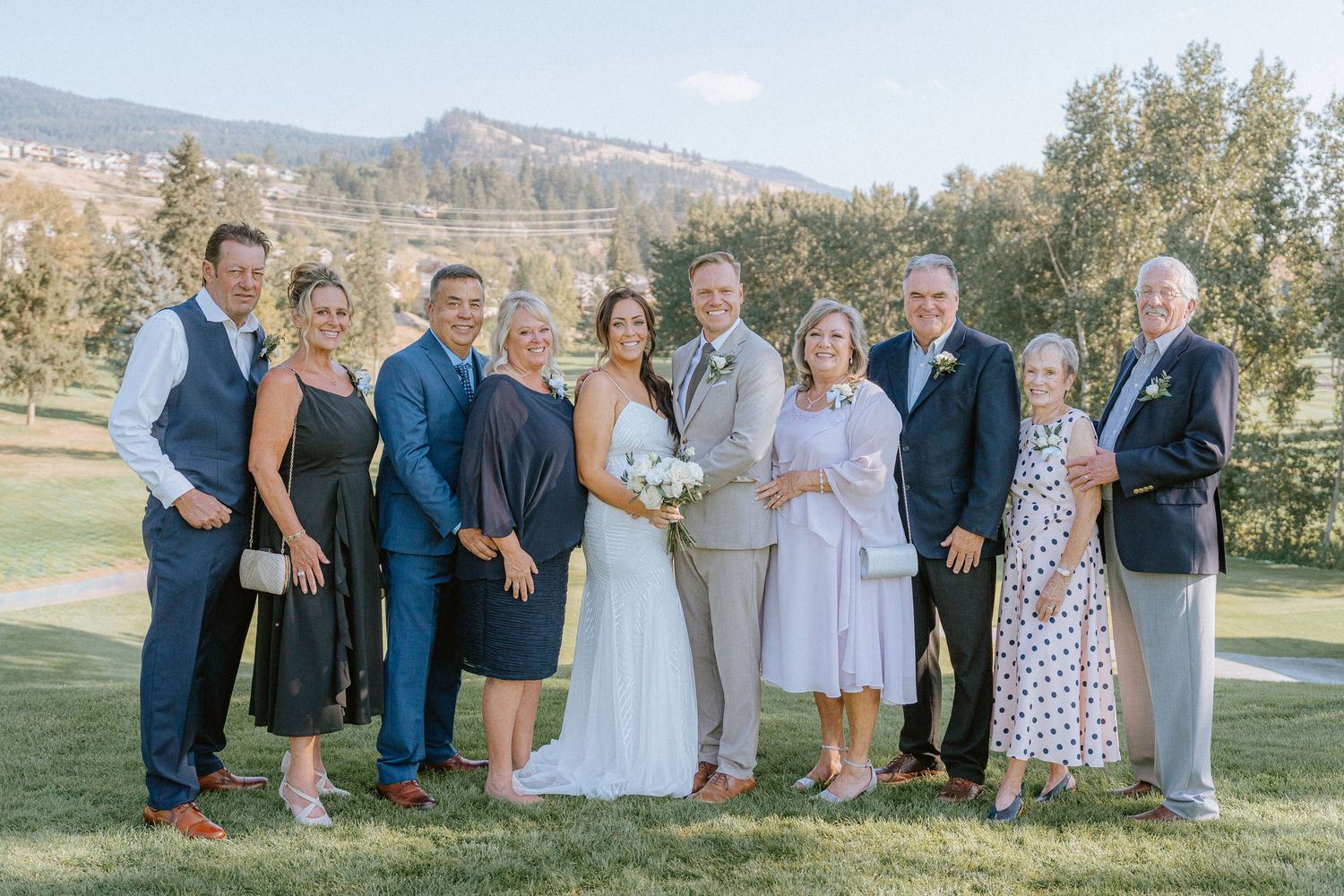 Wedding party gathers for group photos on a grassy hillside overlooking a scenic mountain landscape.