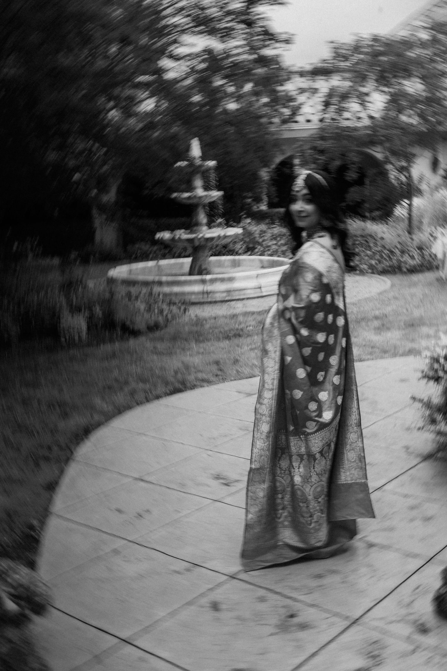 A vintage black and white photograph of someone in a traditional kimono standing near a garden fountain.