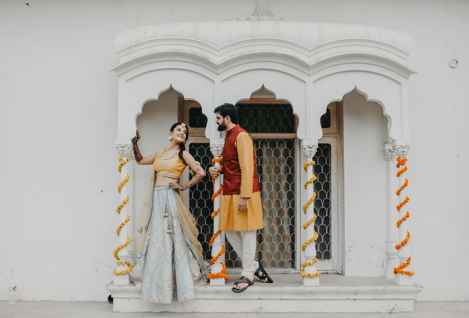 A couple in traditional Indian fusion attire stand in an ornate white archway decorated with orange spiral ribbons.